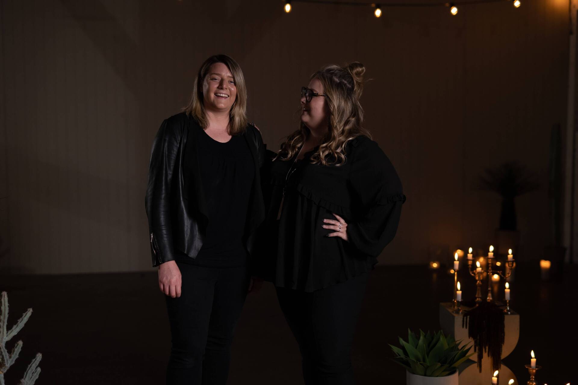 Two women in black clothing smile, posing in a dimly lit space with candles and string lights.