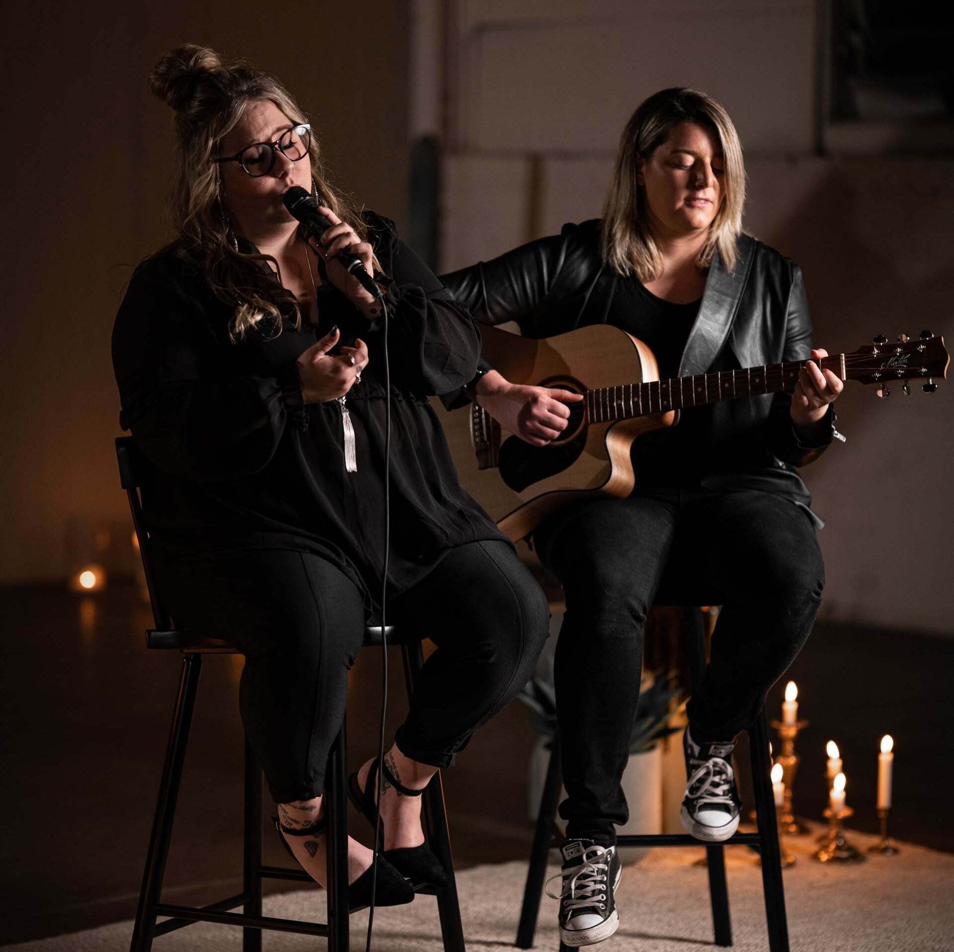 Two women perform acoustic music: one sings into a mic, the other plays guitar. Dim lighting, candles in background.