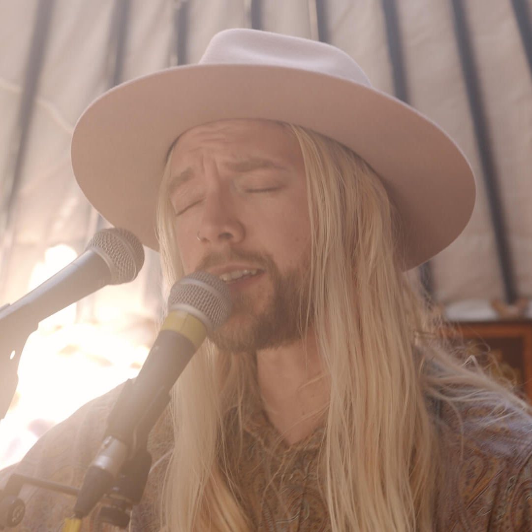 Man with long blonde hair singing into a microphone, wearing a tan hat.