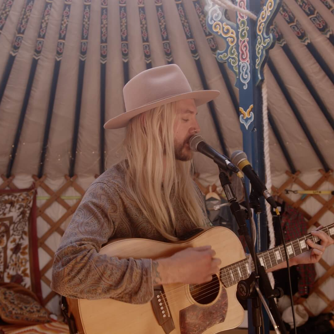 Blonde-haired man wearing a hat, playing guitar and singing into a microphone inside a yurt.