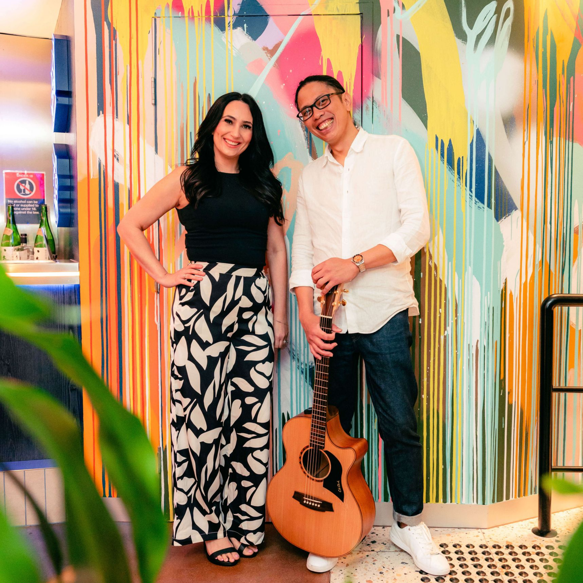 Woman and man posing in front of a colorful wall; the man holds a guitar.