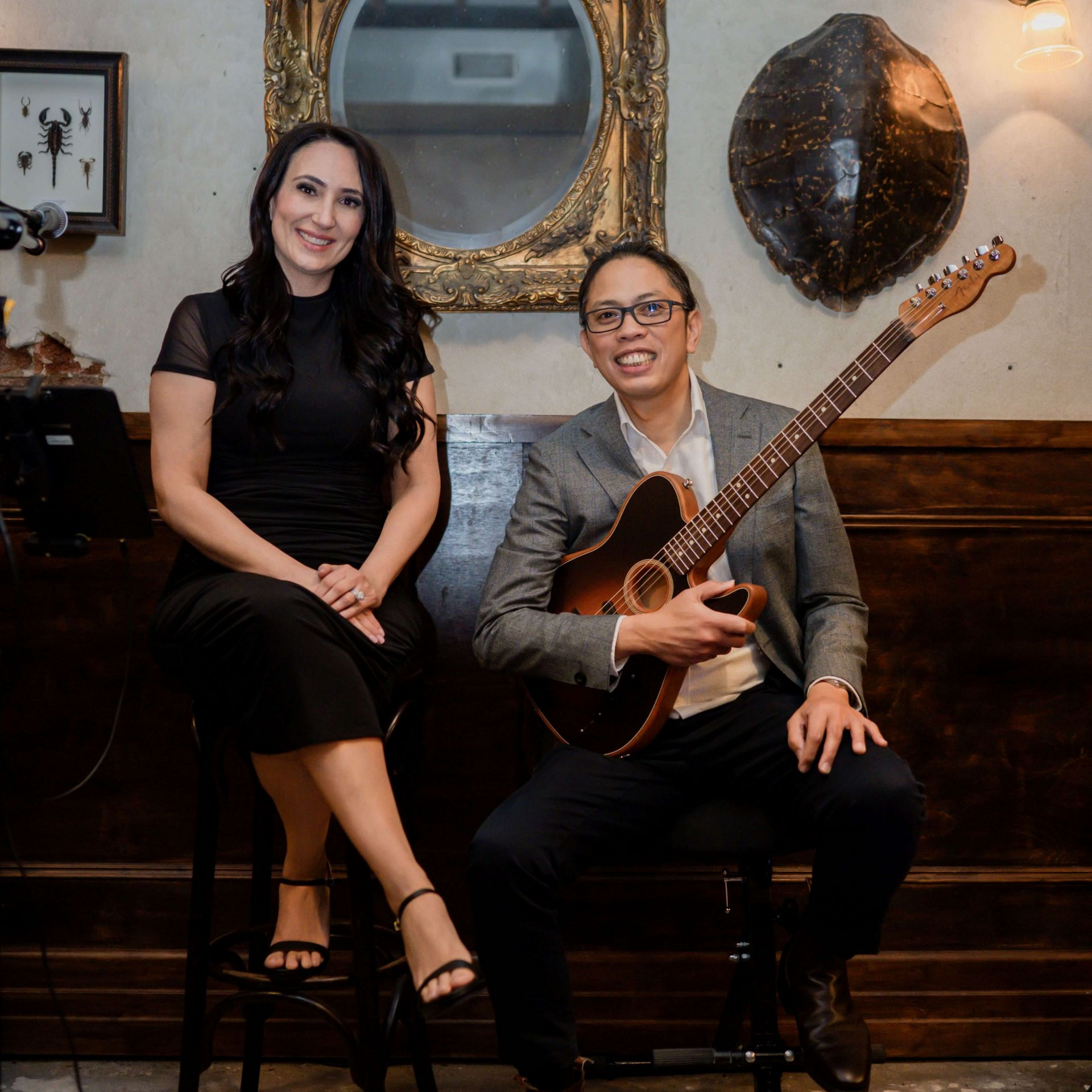 A woman and a man pose in a room. The man holds a guitar. They sit on stools.