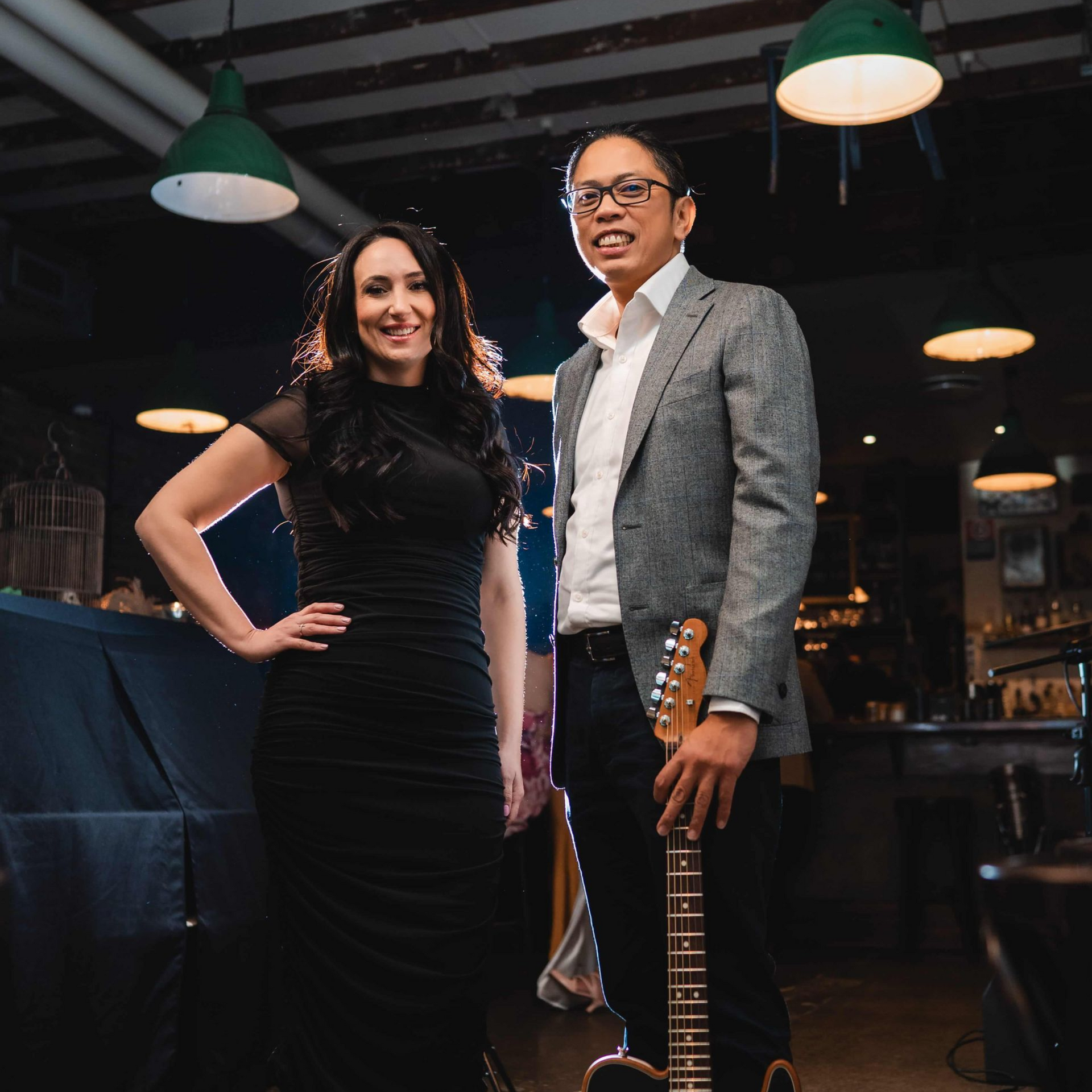 A woman in a black dress and a man in a gray blazer stand in a dimly lit bar. The man holds a guitar.