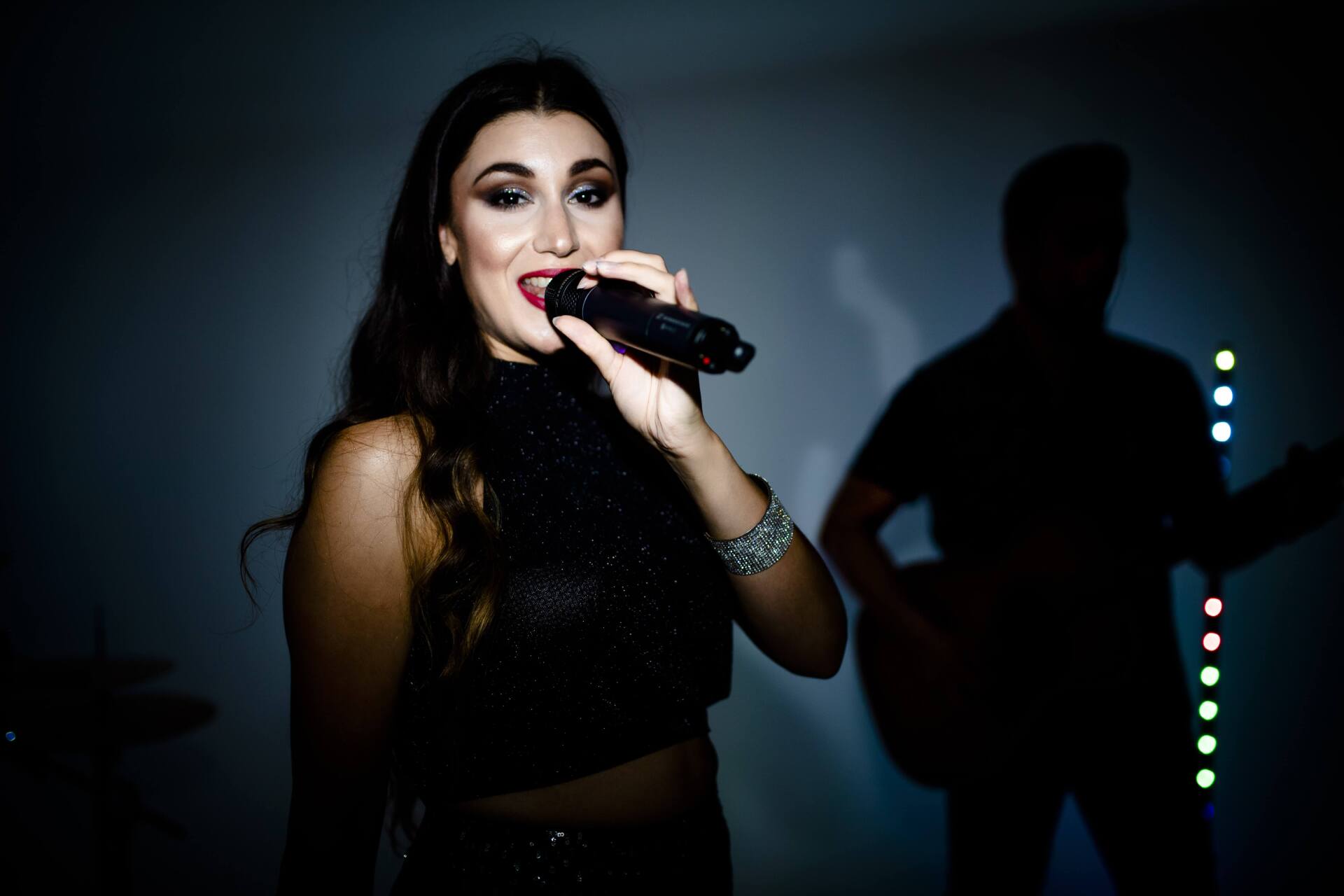 Woman singing into a microphone with a bandmate silhouetted in the background. Dark, moody lighting.