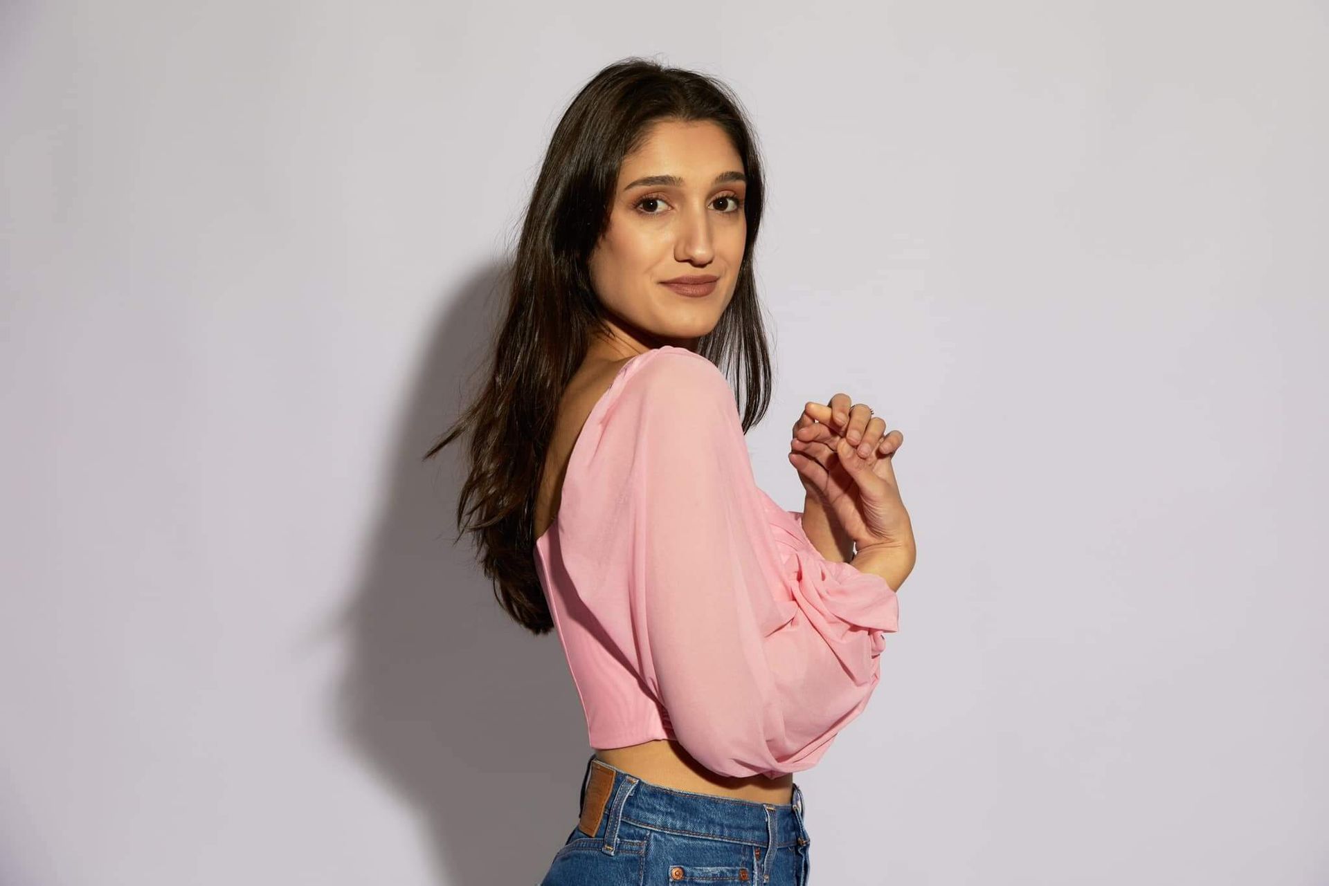 Woman in pink top and jeans, hands clasped, looking at the viewer with a slight smile against a white backdrop.