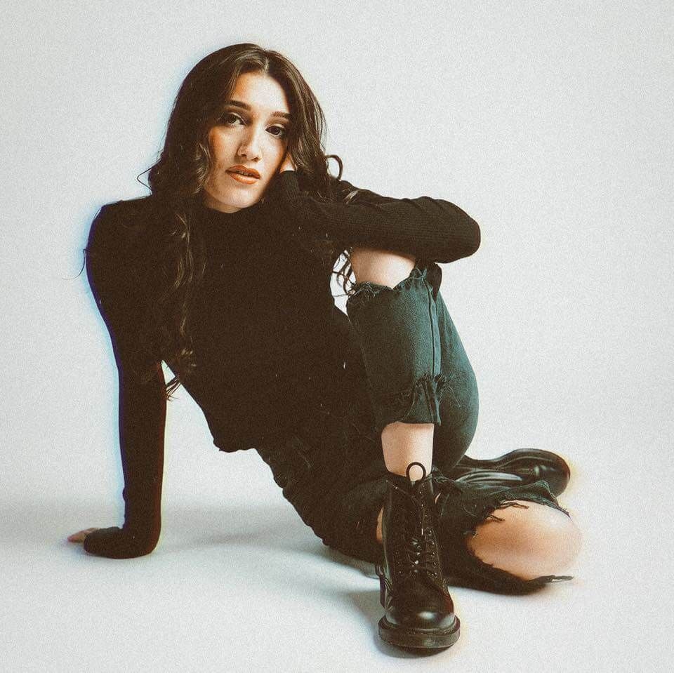 Woman in black top and ripped jeans, boots, sitting with a serious expression. Light background.