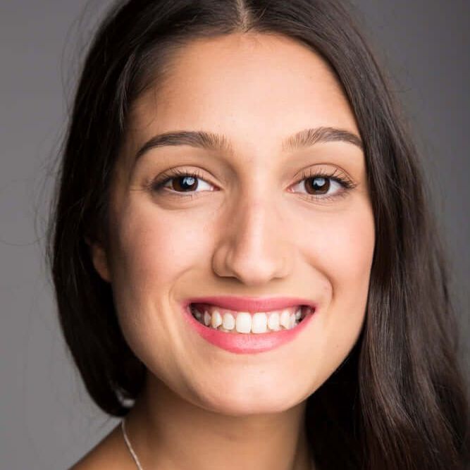 Woman with dark hair smiles at the camera, wearing a silver necklace, against a gray background.
