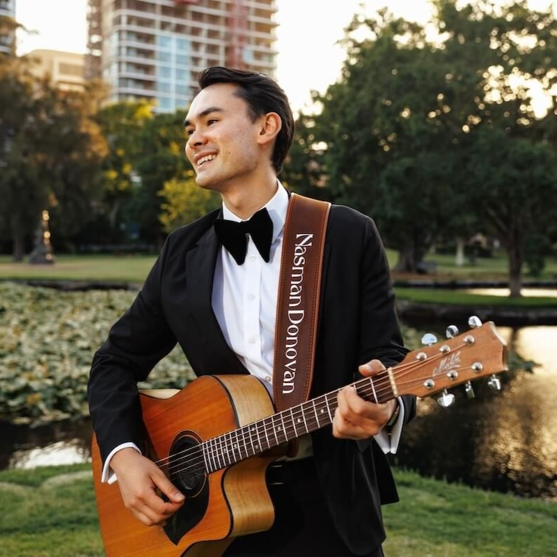 Man in a black suit playing acoustic guitar outdoors near a pond and city buildings