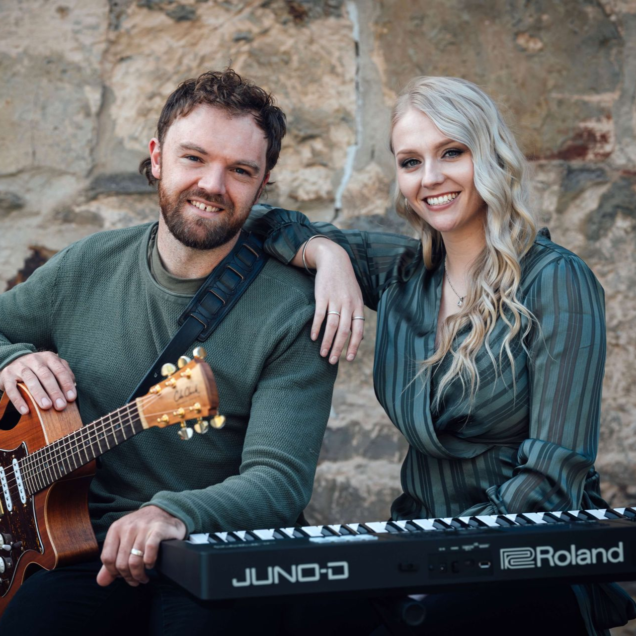 A musical duo with a guitar and keyboard smiling, sitting in front of a stone wall.