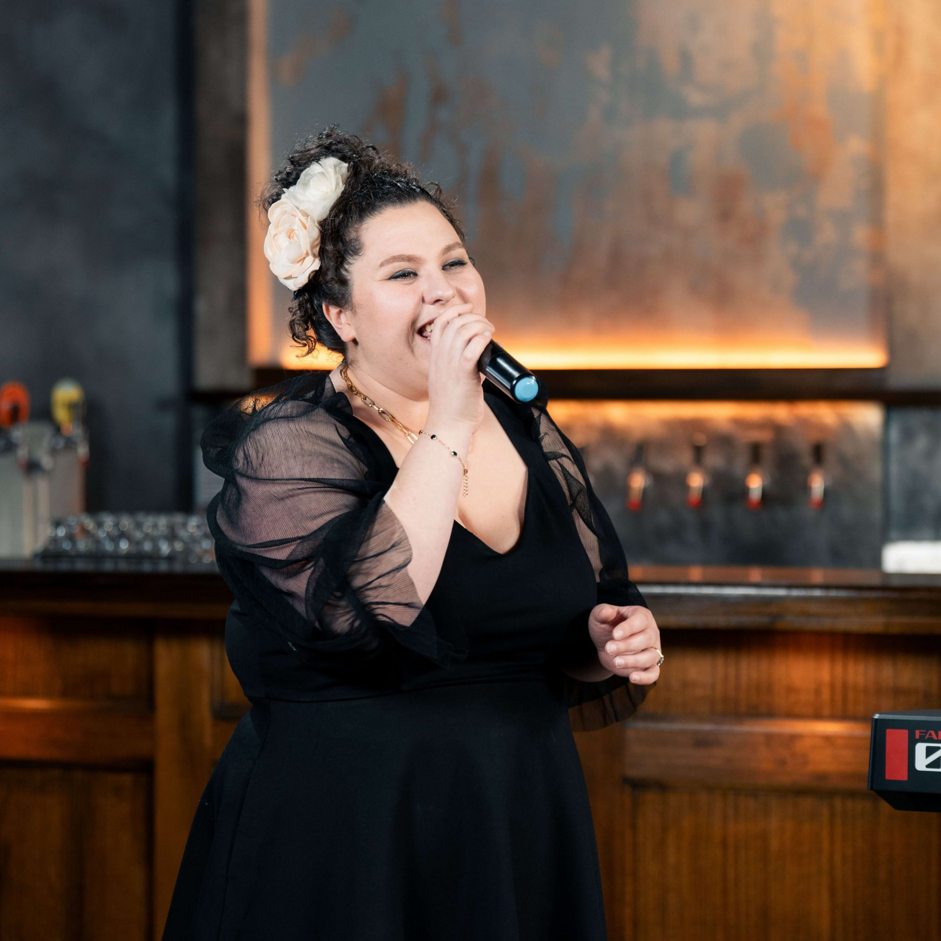 Woman singing into a microphone, wearing a black dress with flower in her hair, at a bar.