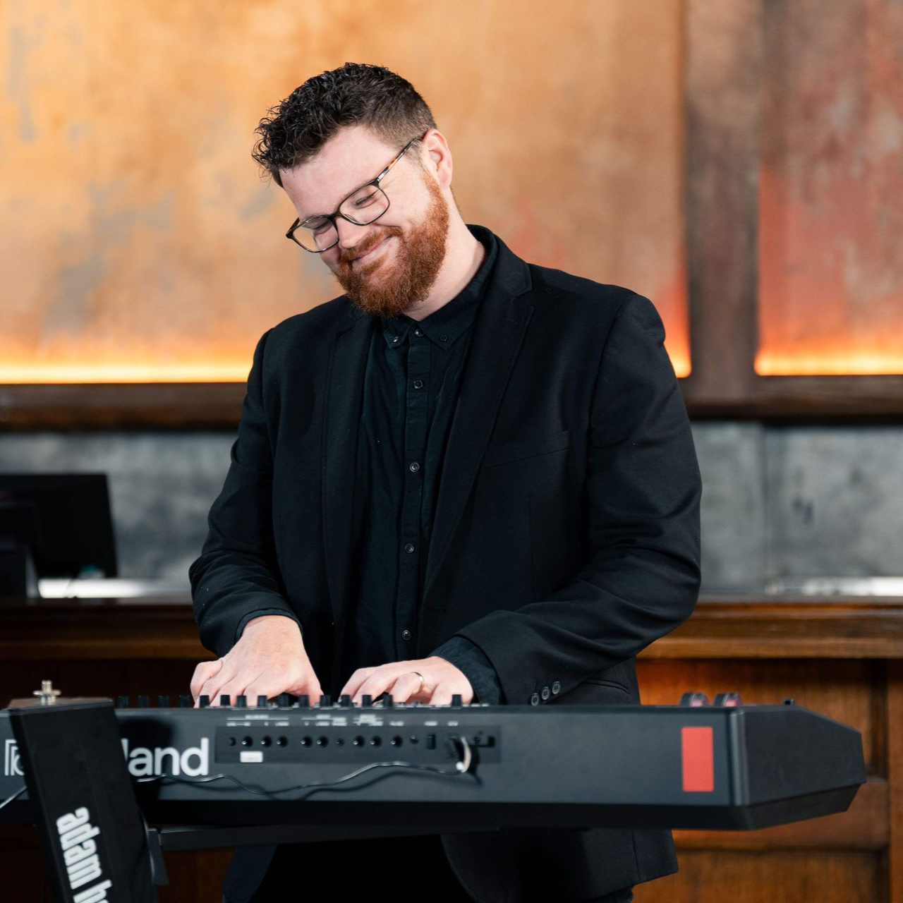 Man with glasses playing a black keyboard, smiling. He's wearing a black suit in a room with orange walls.