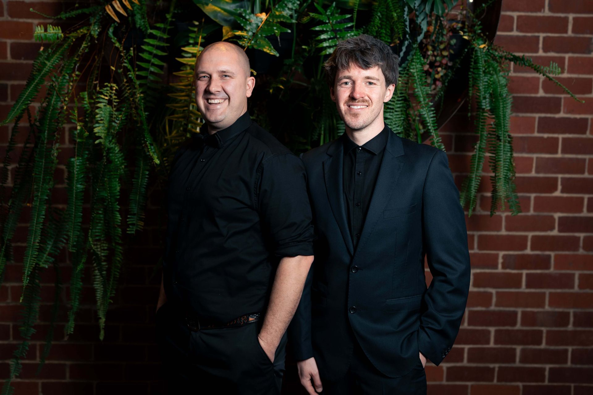 Two men in black outfits pose in front of a brick wall and greenery. One smiles.