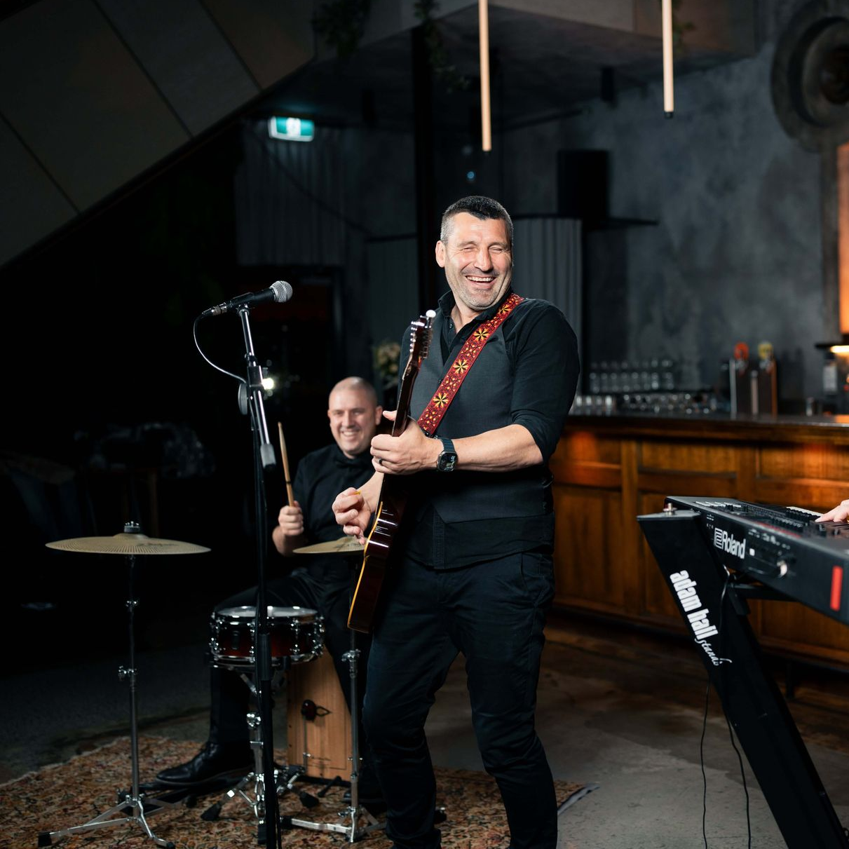 Band playing live music. Guitarist smiling, drummer in background, keyboard and dim-lit bar setting.