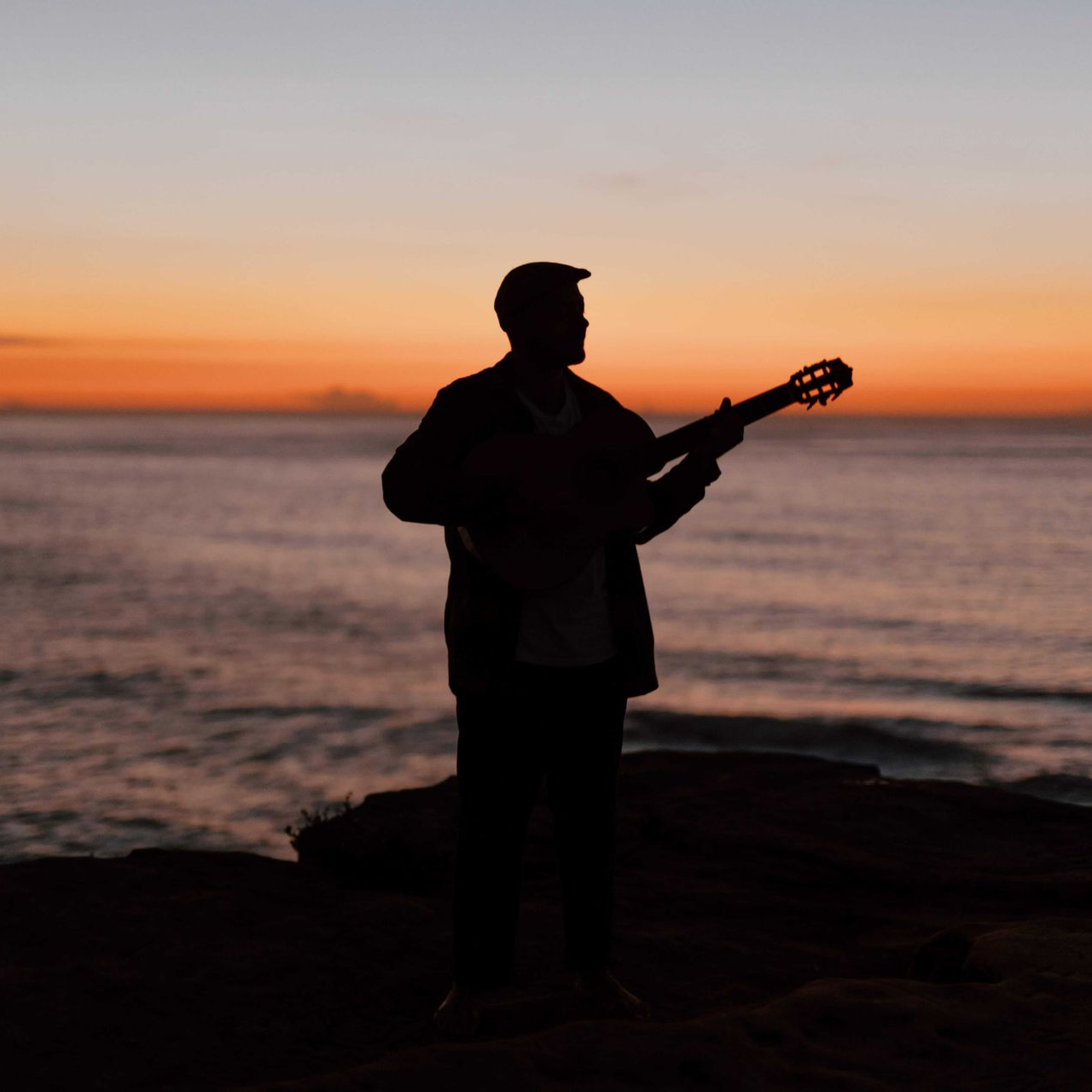 Silhouette of a person playing a guitar at the beach during a colorful sunset over the ocean.