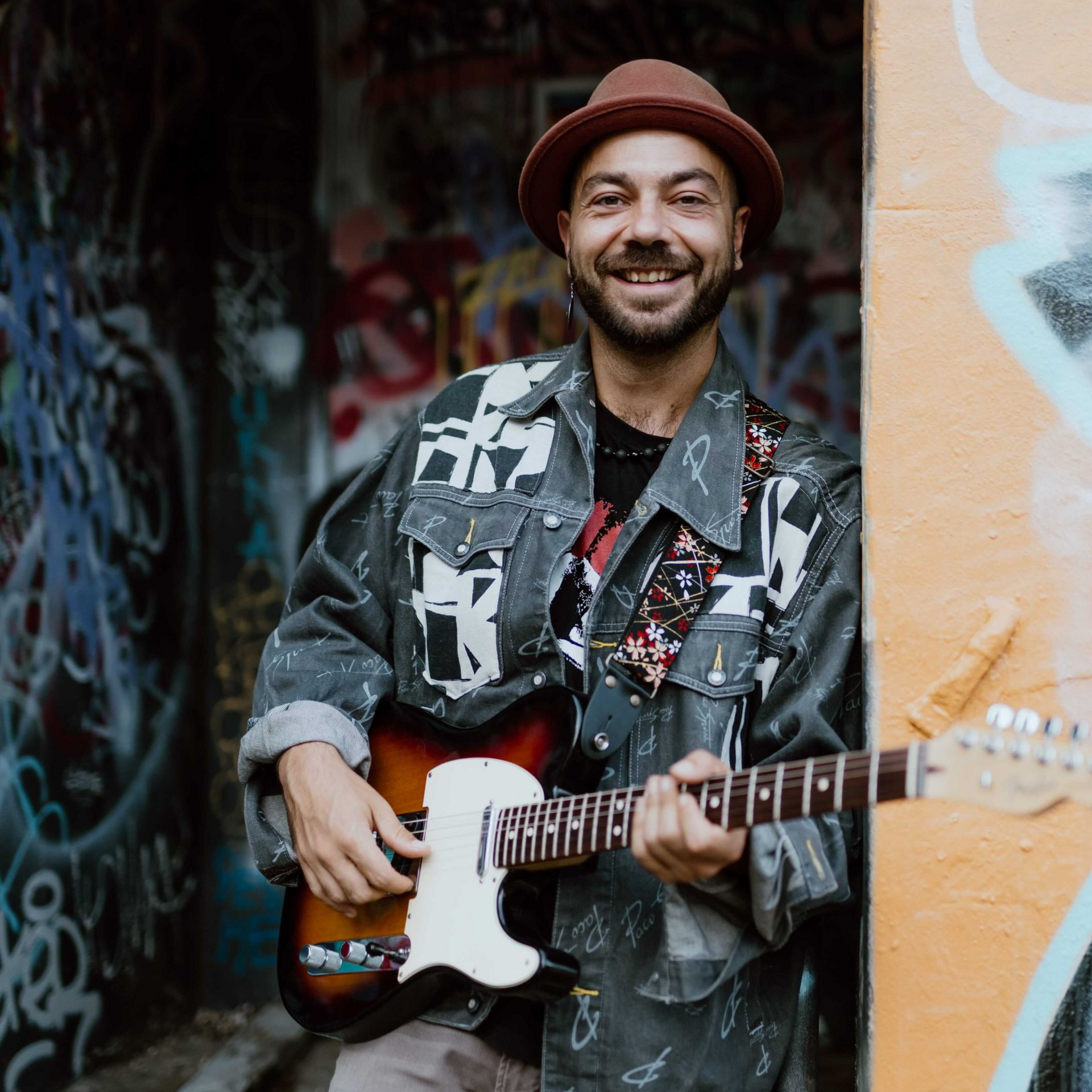 Smiling musician holding guitar, wearing a hat and jacket with colorful graffiti in background.