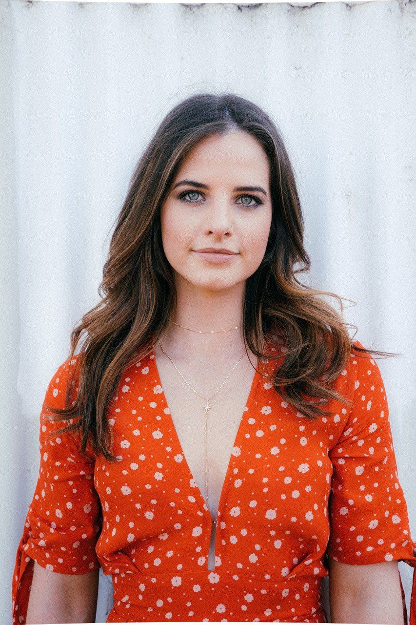 A woman in a red dress with white polka dots is standing in front of a white wall.