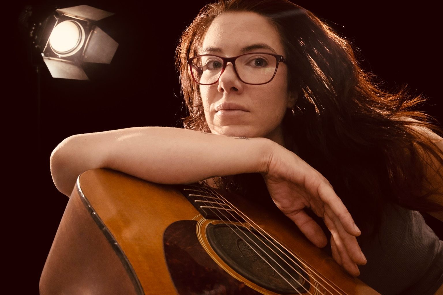 Woman with glasses, leaning on acoustic guitar, lit by a spotlight against a dark background.