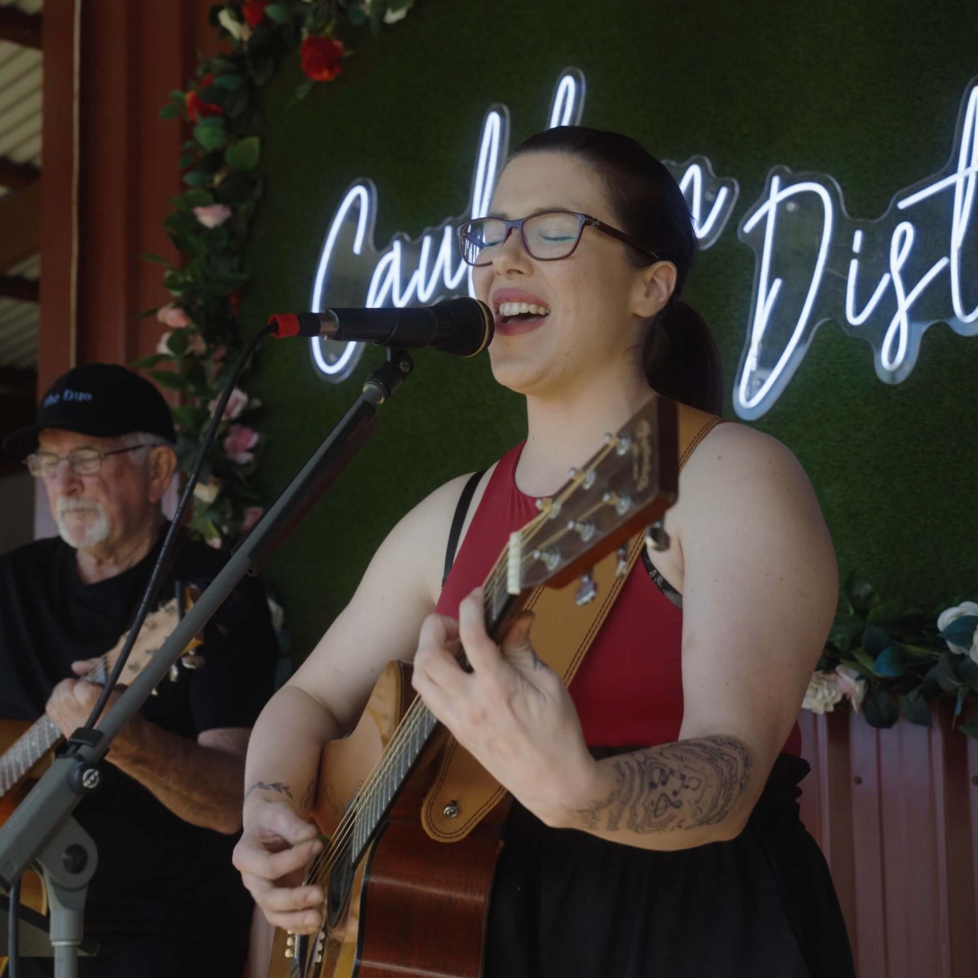Woman singing and playing acoustic guitar onstage, with another guitarist in the background.