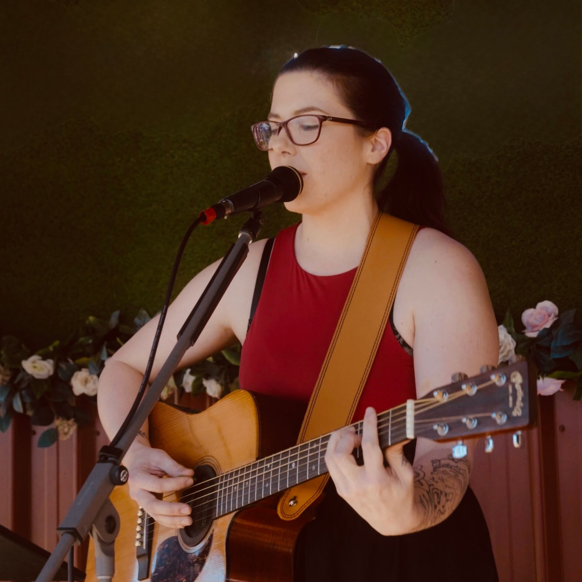 Woman playing acoustic guitar and singing into a microphone outdoors. She wears glasses, a red top, and a wide guitar strap.