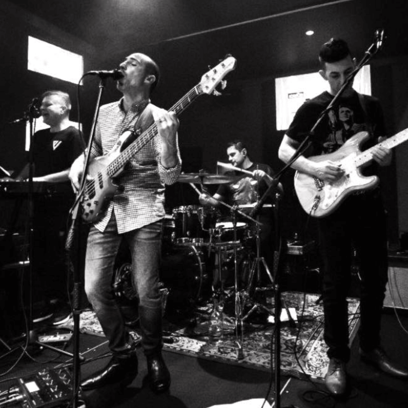 Band playing on stage: a bassist sings into a mic, guitarist, drummer, and keyboardist performing.