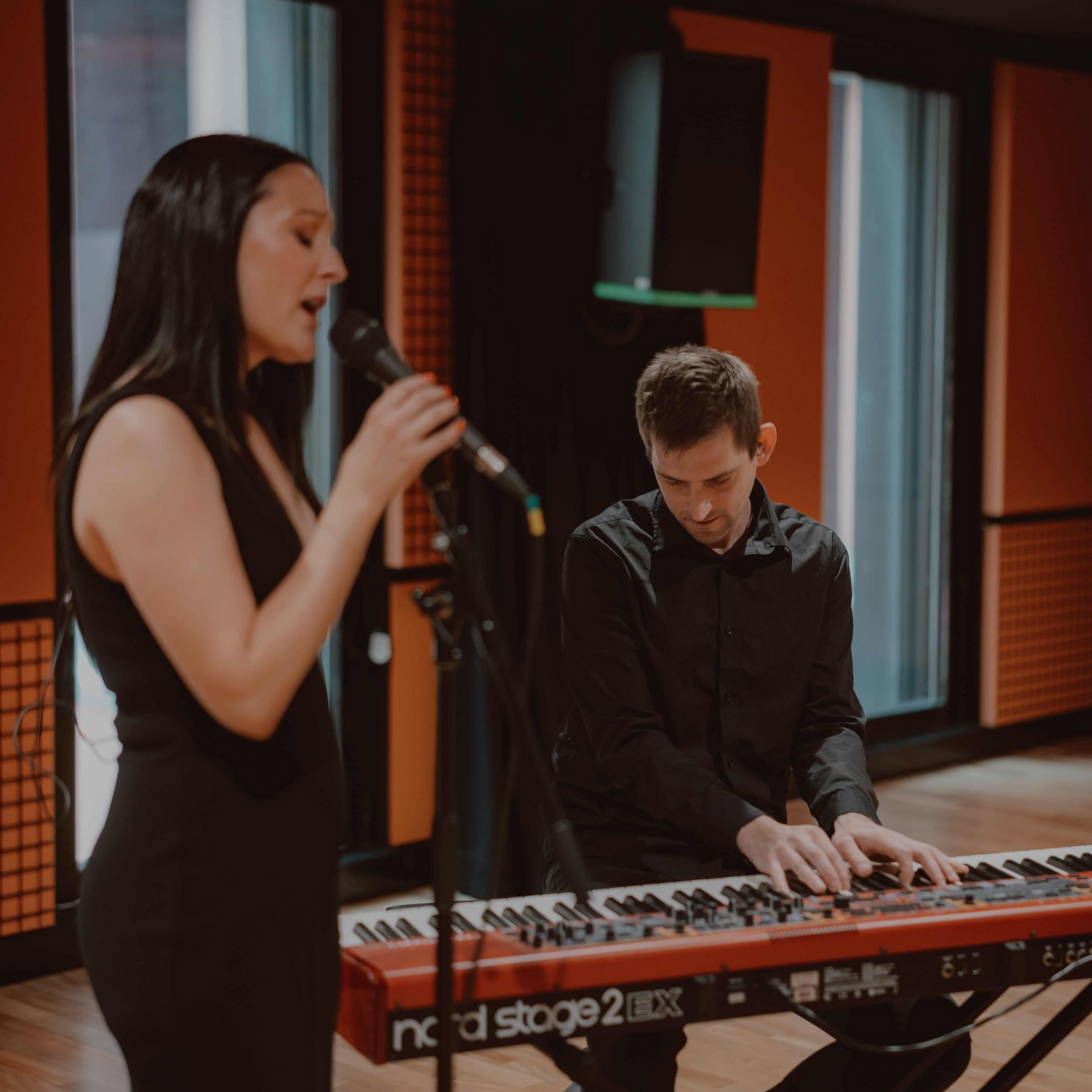 Woman singing into a microphone, man playing a red keyboard in a studio.