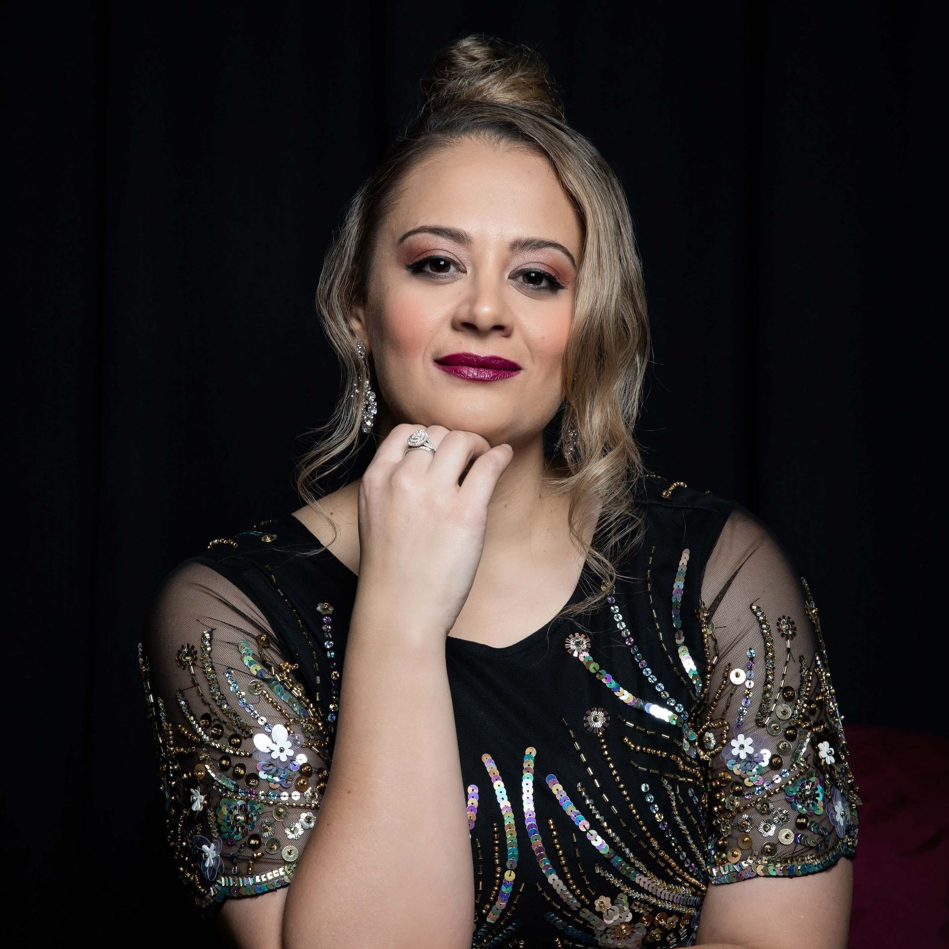 Woman in black dress with beading, hand on chin, dark lipstick, looking at camera.