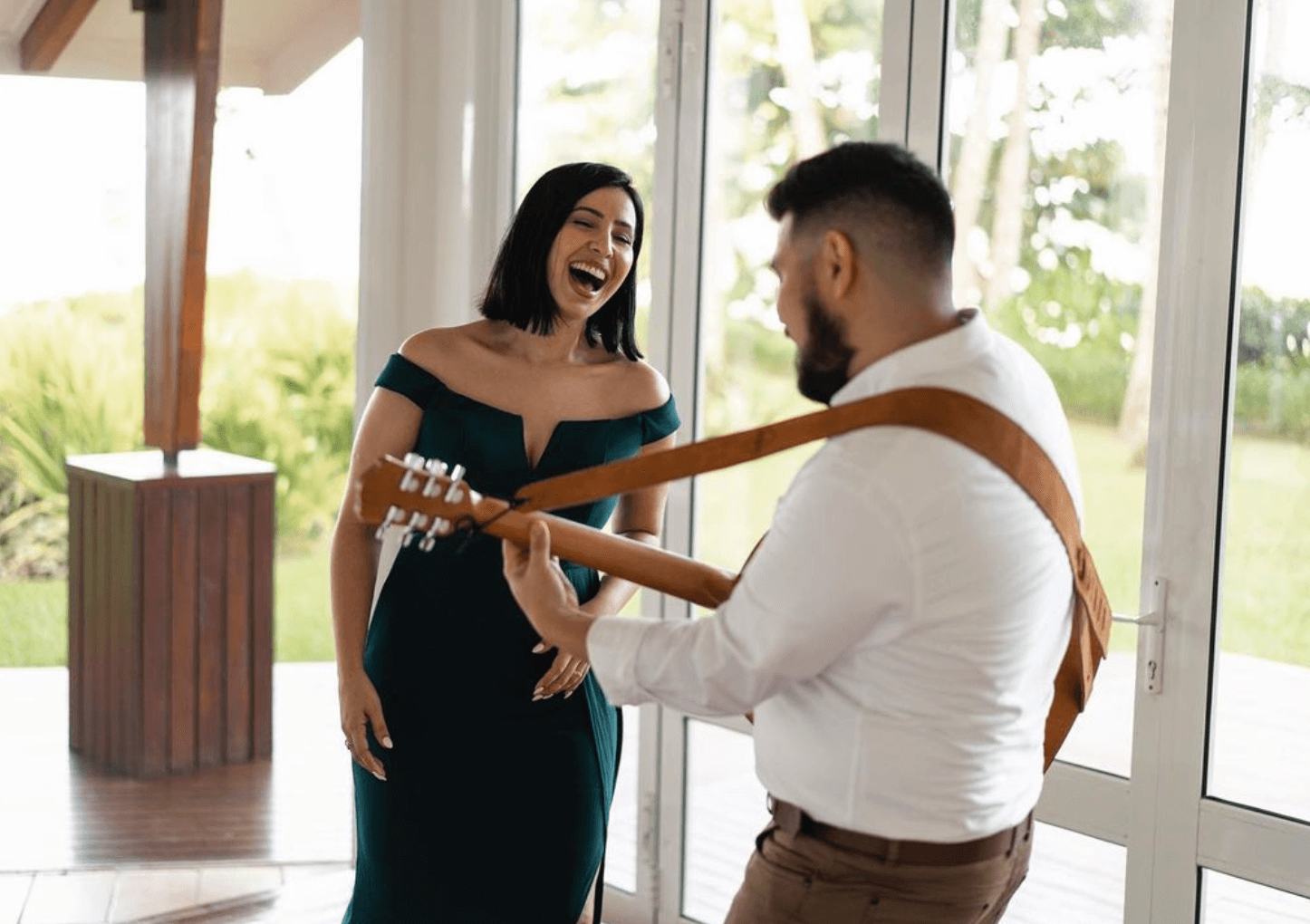 Man serenading a smiling woman with a guitar in a bright, modern room by large windows