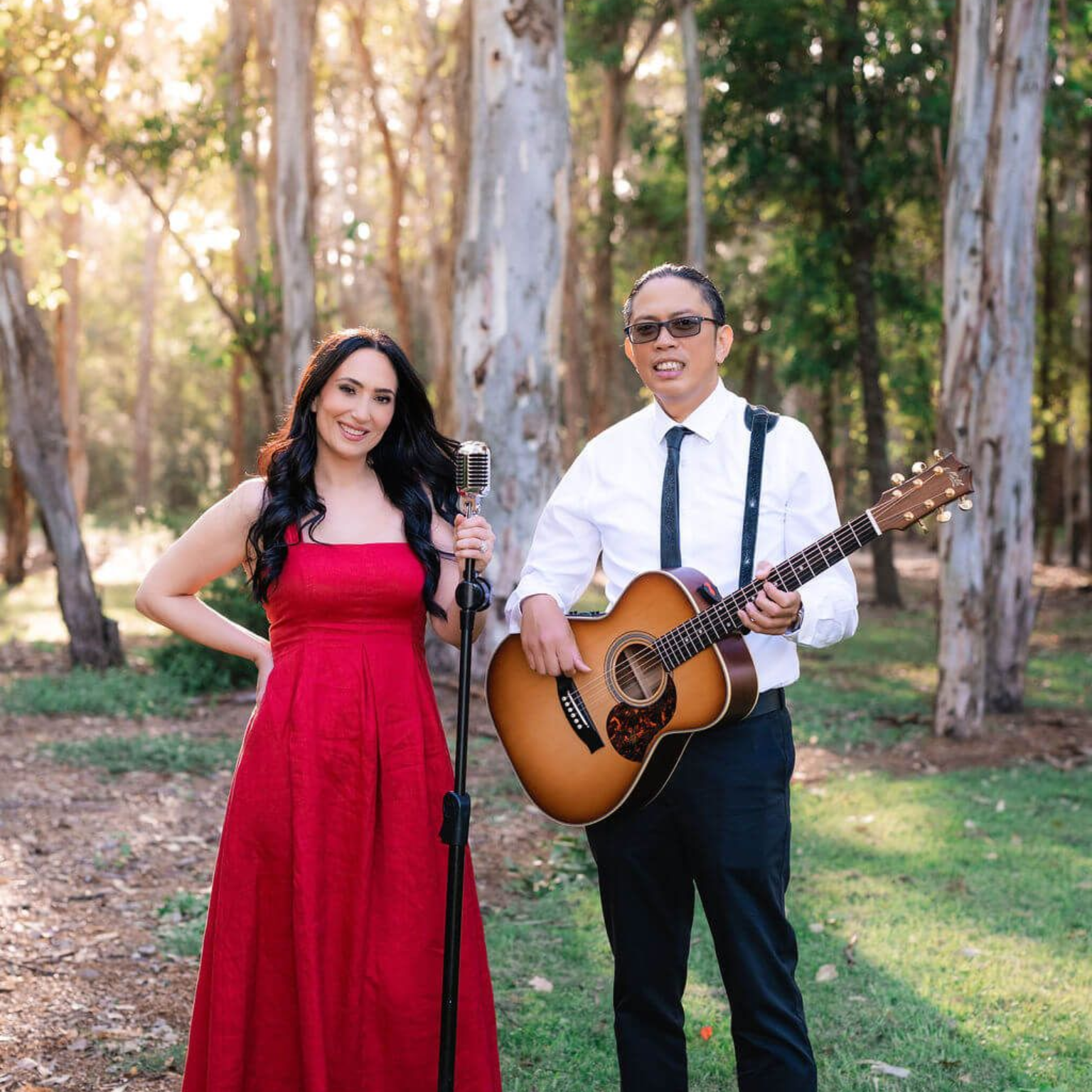 A person in a red dress stands by a microphone next to a person in a white shirt playing an acoustic guitar in a forest.