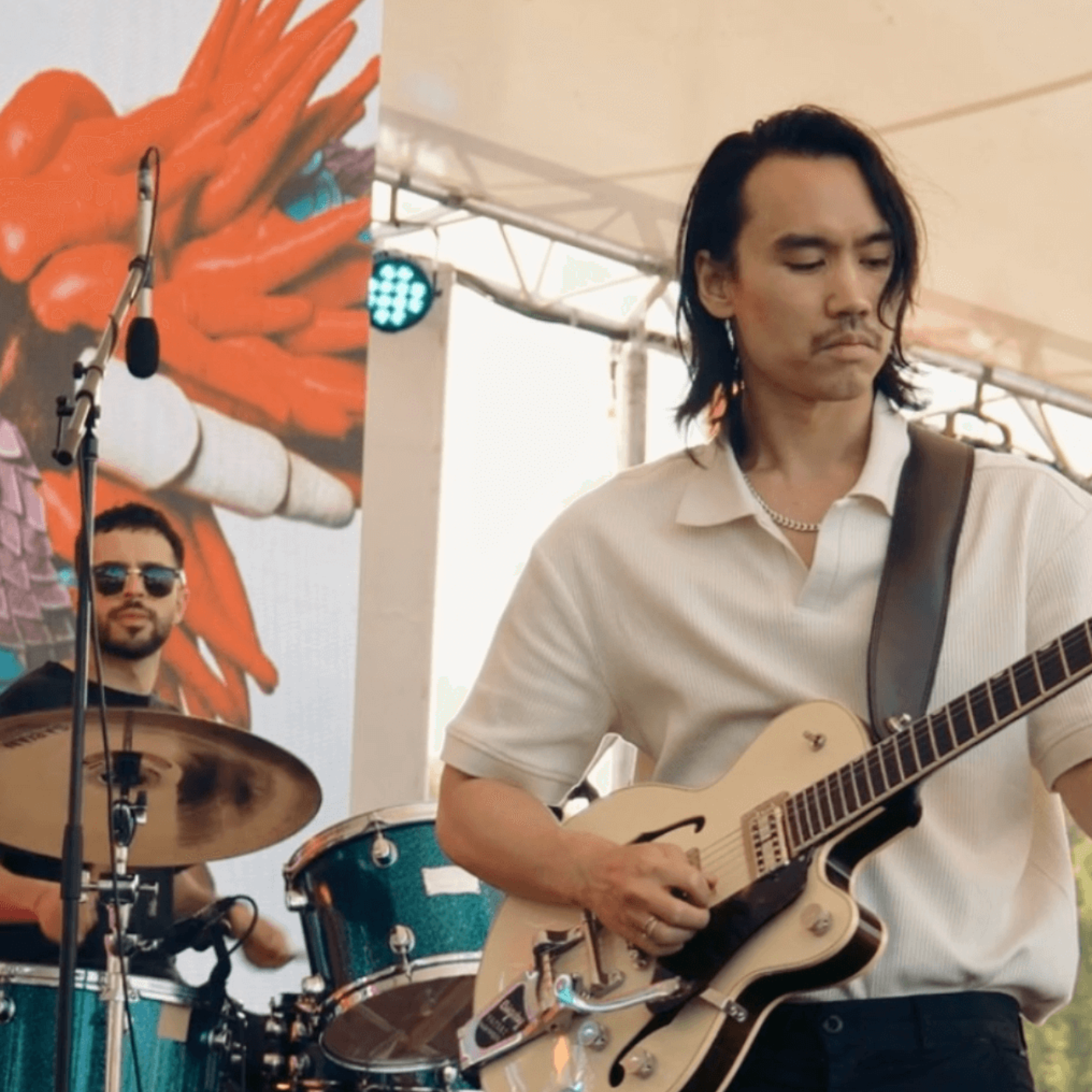 A guitarist playing on stage with a drummer in the background. The guitarist is wearing a cream shirt.