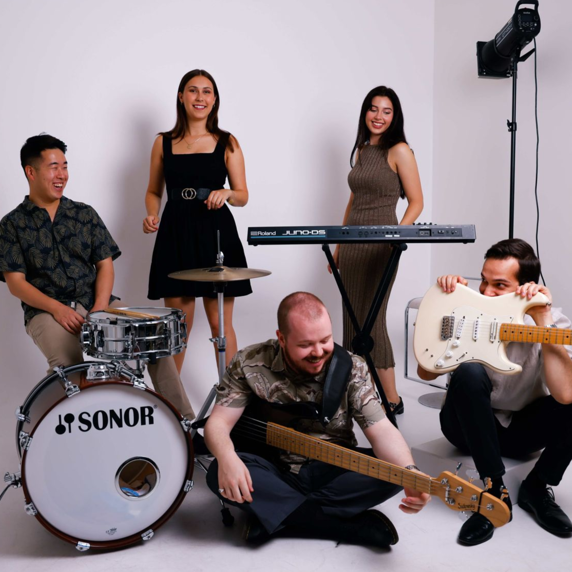 Band poses in a white studio: drummer, keyboardist, guitarist, two women stand behind.