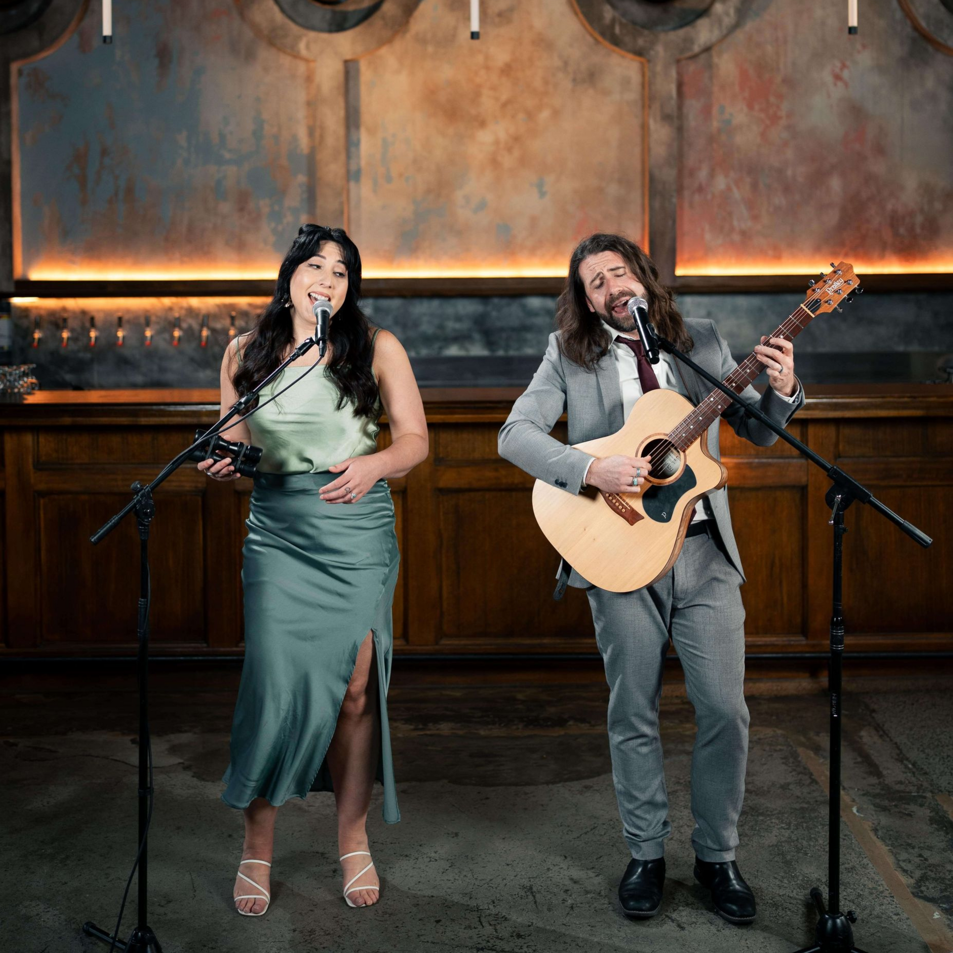 Woman singing, man playing acoustic guitar on stage. They are both in front of a wall with decorative elements.