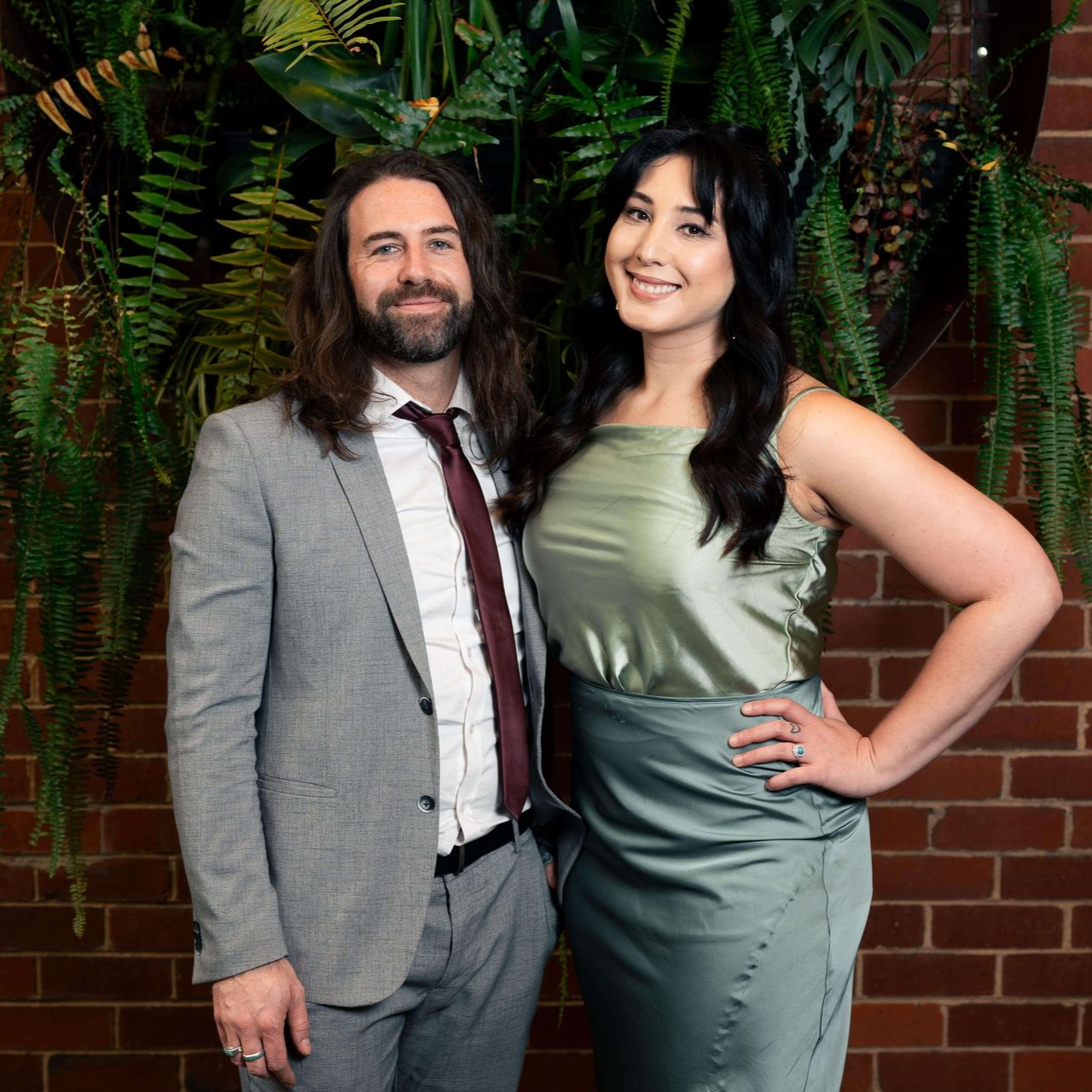 A man and woman pose together. The woman wears a sage dress, the man a suit. Brick wall and greenery in background.