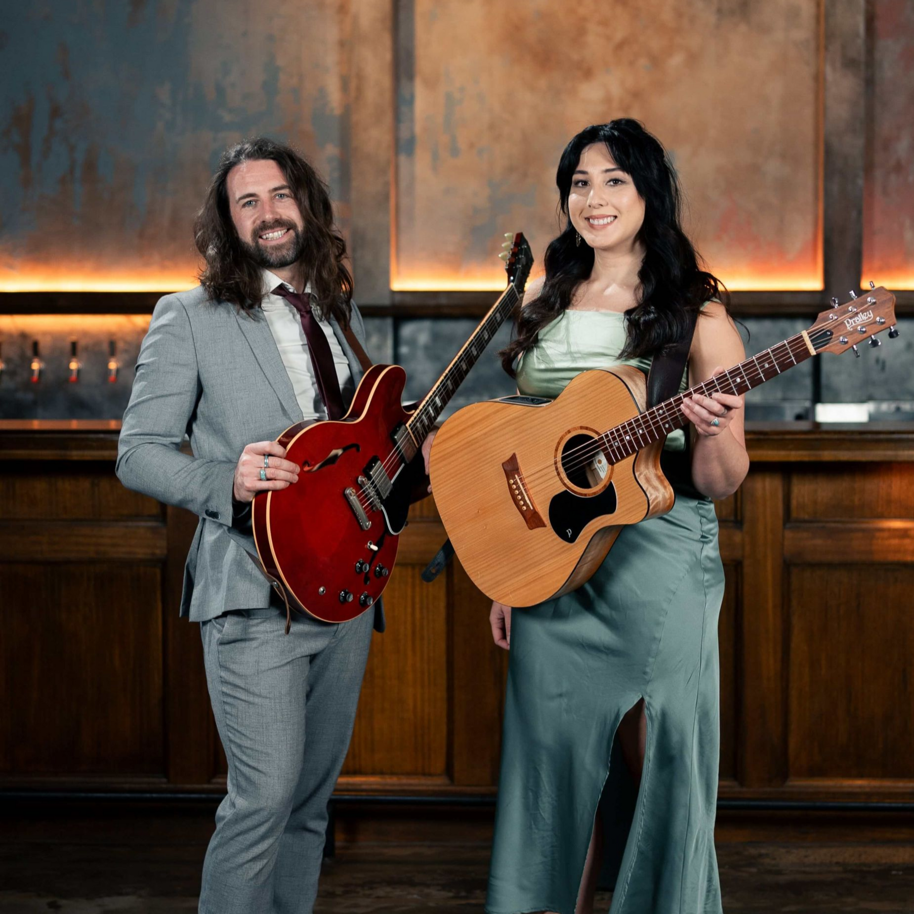 A man and woman with guitars pose indoors. Man in gray suit, woman in green dress, both smiling.