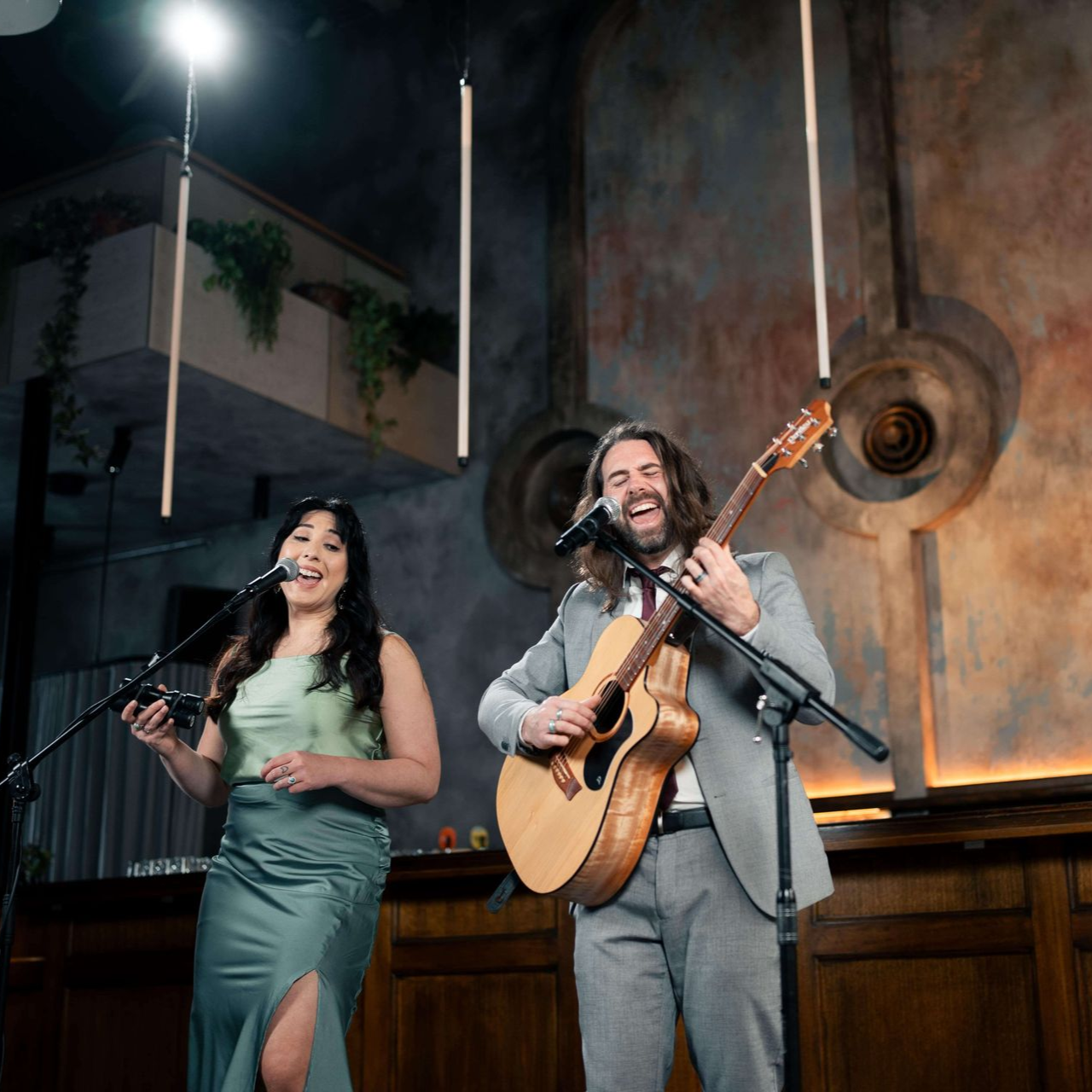 Two musicians performing on stage: woman singing, man playing guitar. Dark, rustic setting.