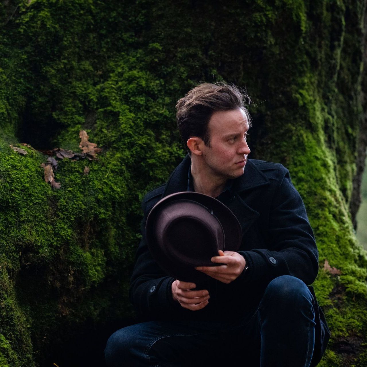 A man is sitting in front of a tree holding a hat.