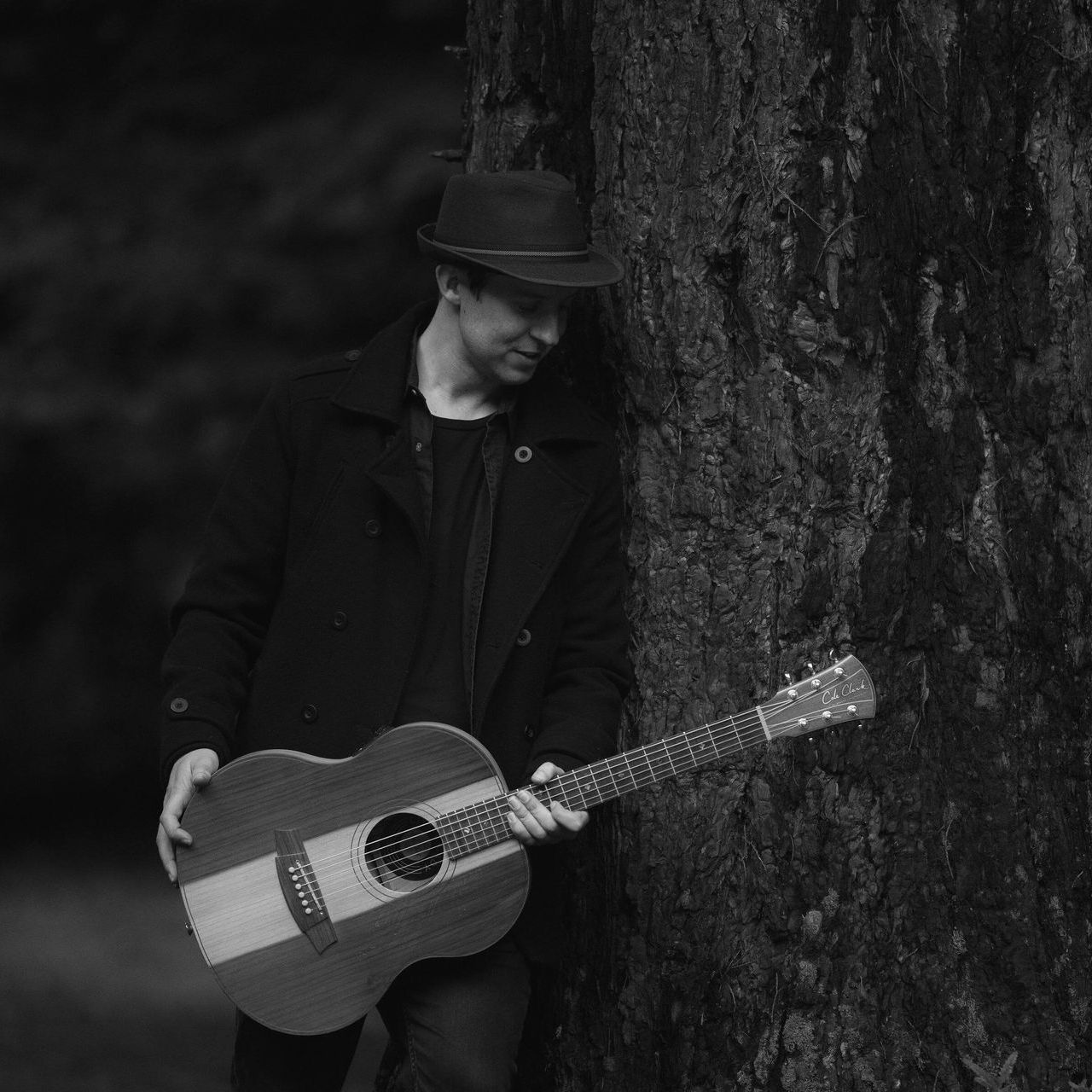A man in a hat is leaning against a tree while holding an acoustic guitar.
