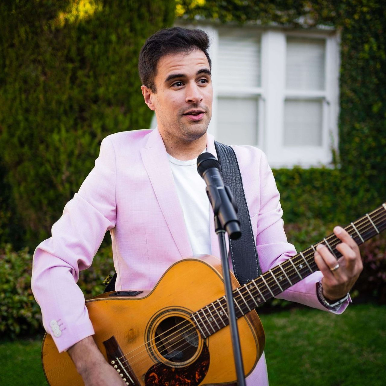A man in a pink jacket is singing into a microphone while holding a guitar