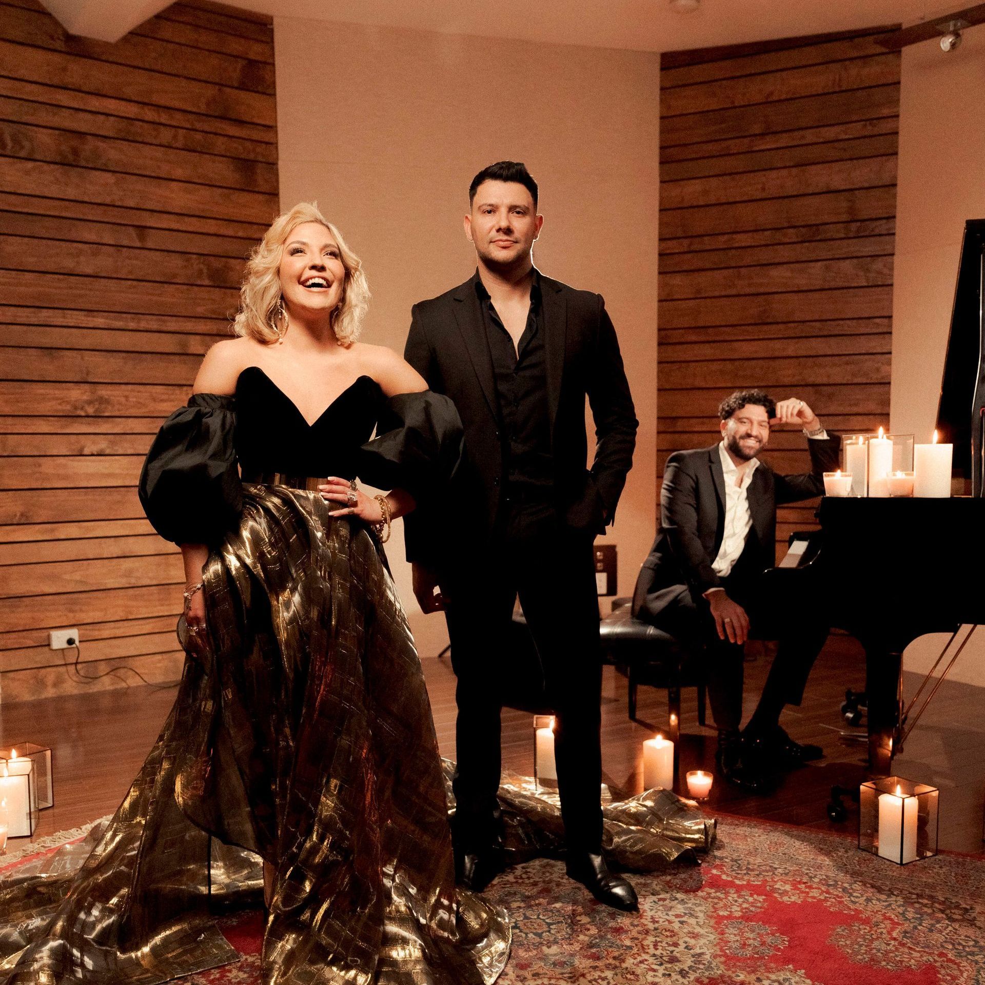 Three people in formal attire pose in a candlelit room: woman in gold dress, two men in black.