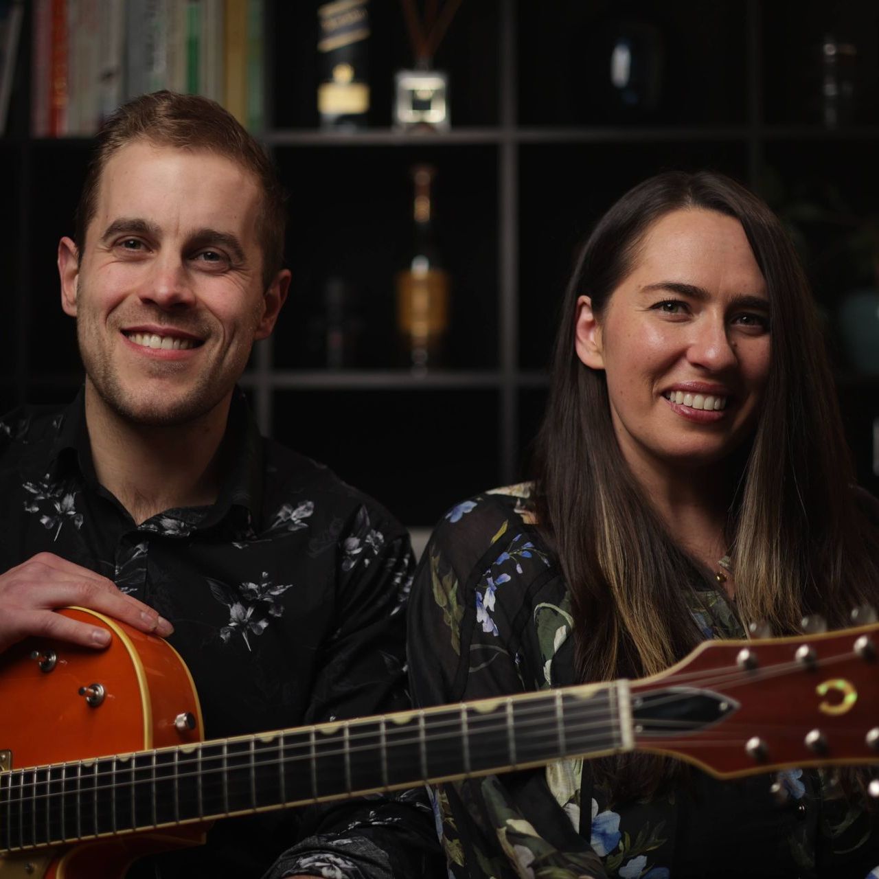 Man and woman smiling, holding guitars. Indoor setting with bookshelves.