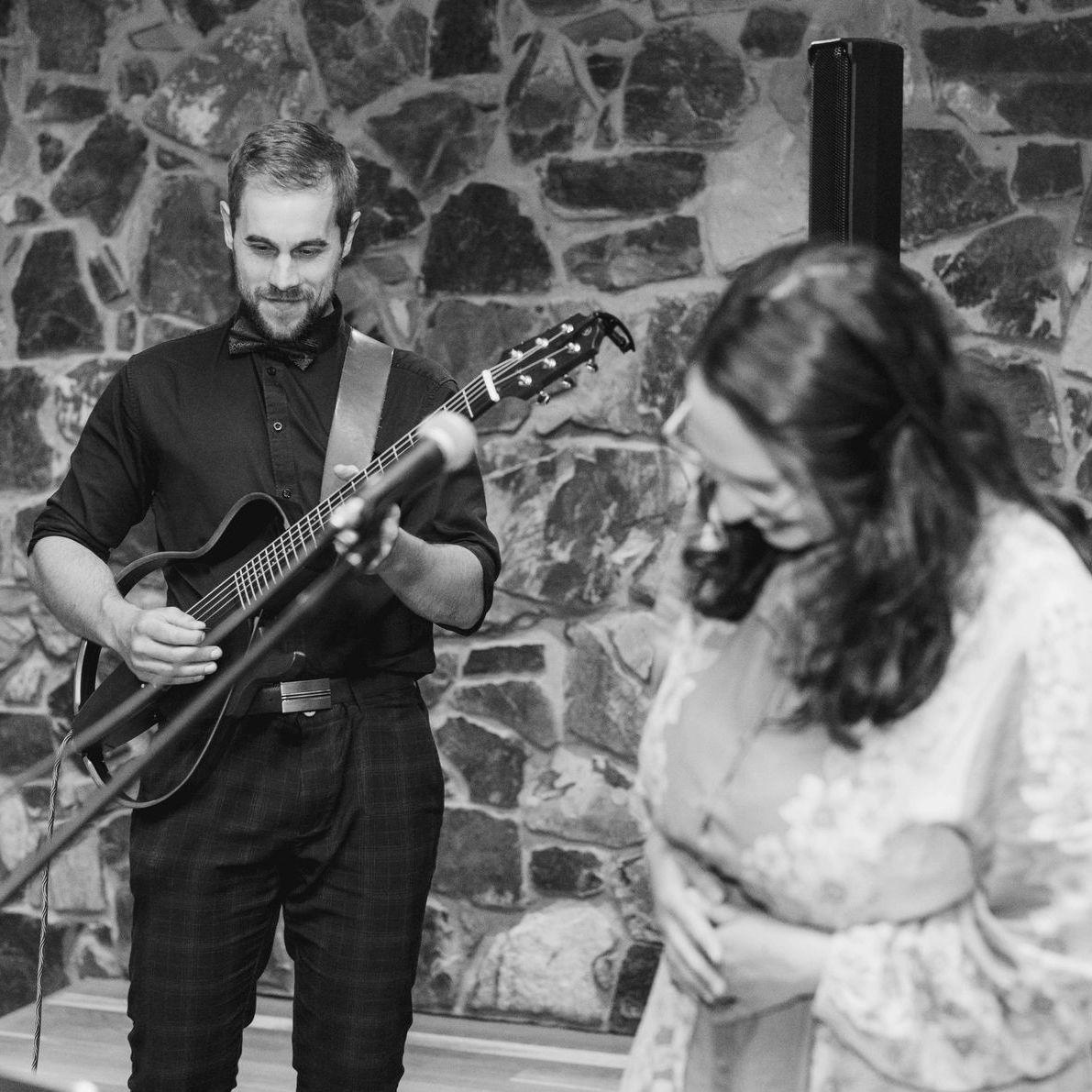 Man plays guitar and sings into a microphone, woman sings nearby, both on a stage with a stone wall backdrop.