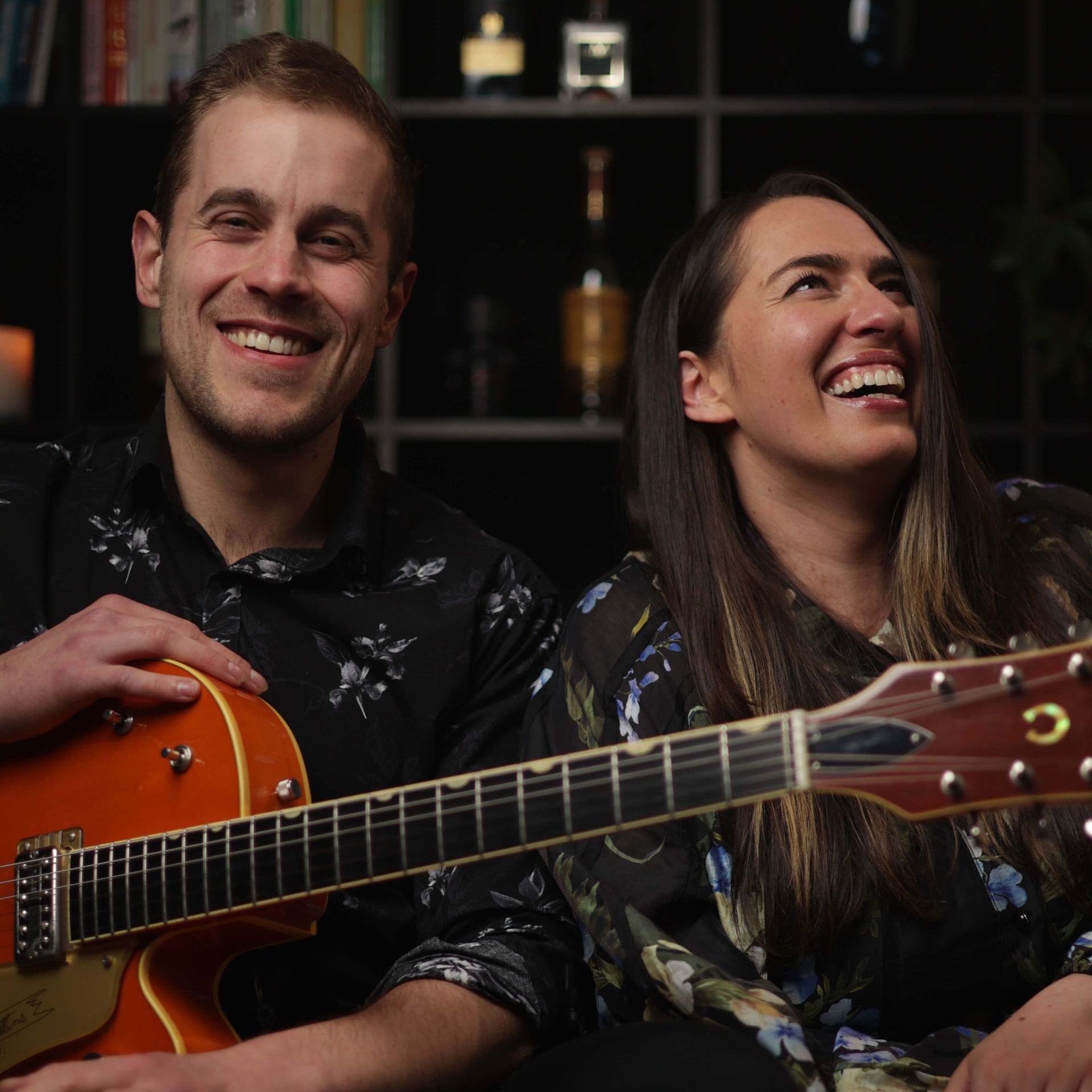 Man and woman smile, holding a guitar, in a cozy setting.