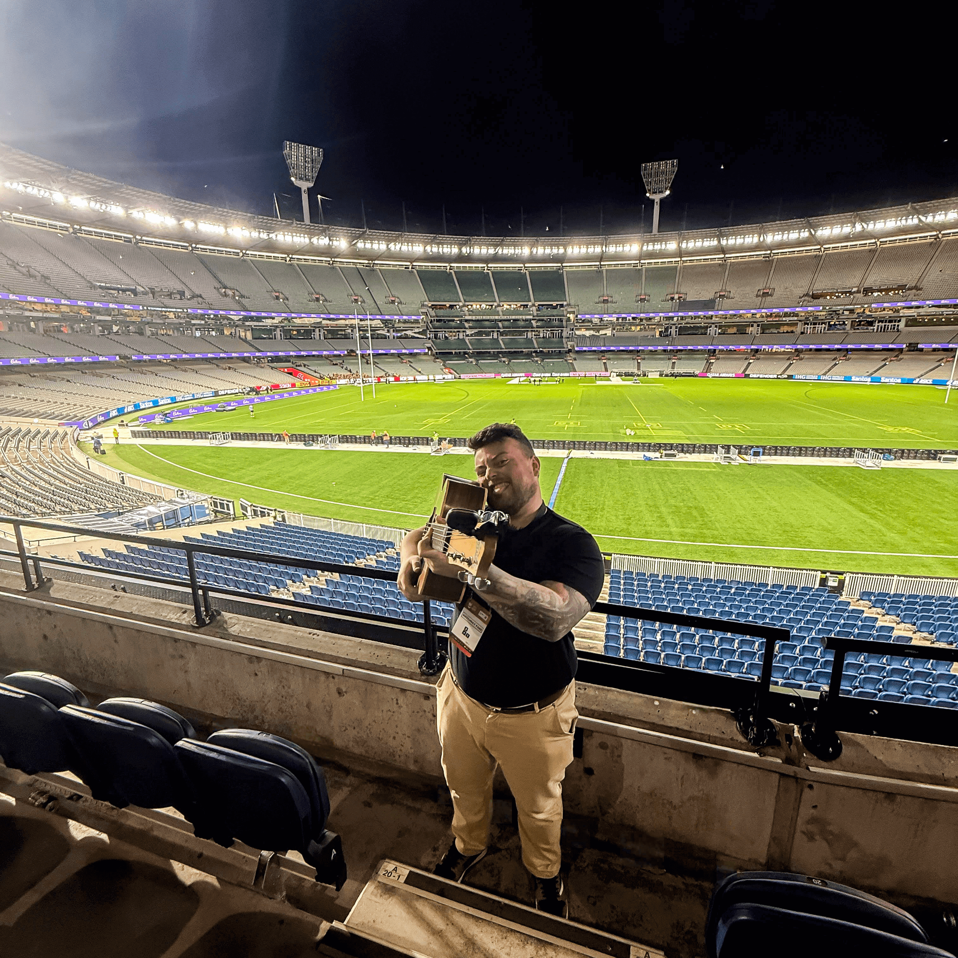 Man in stadium holding a trophy, night. Beige pants, black shirt, and a stadium with green field.