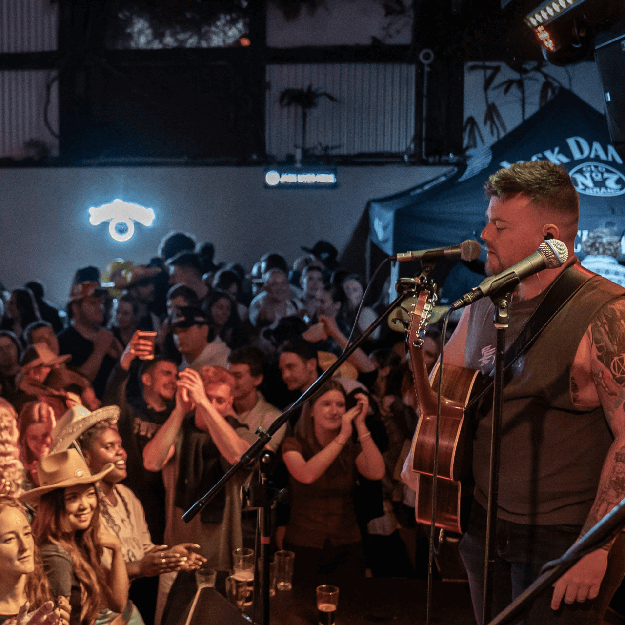 A musician plays guitar and sings on stage in a crowded bar. People cheer and clap.