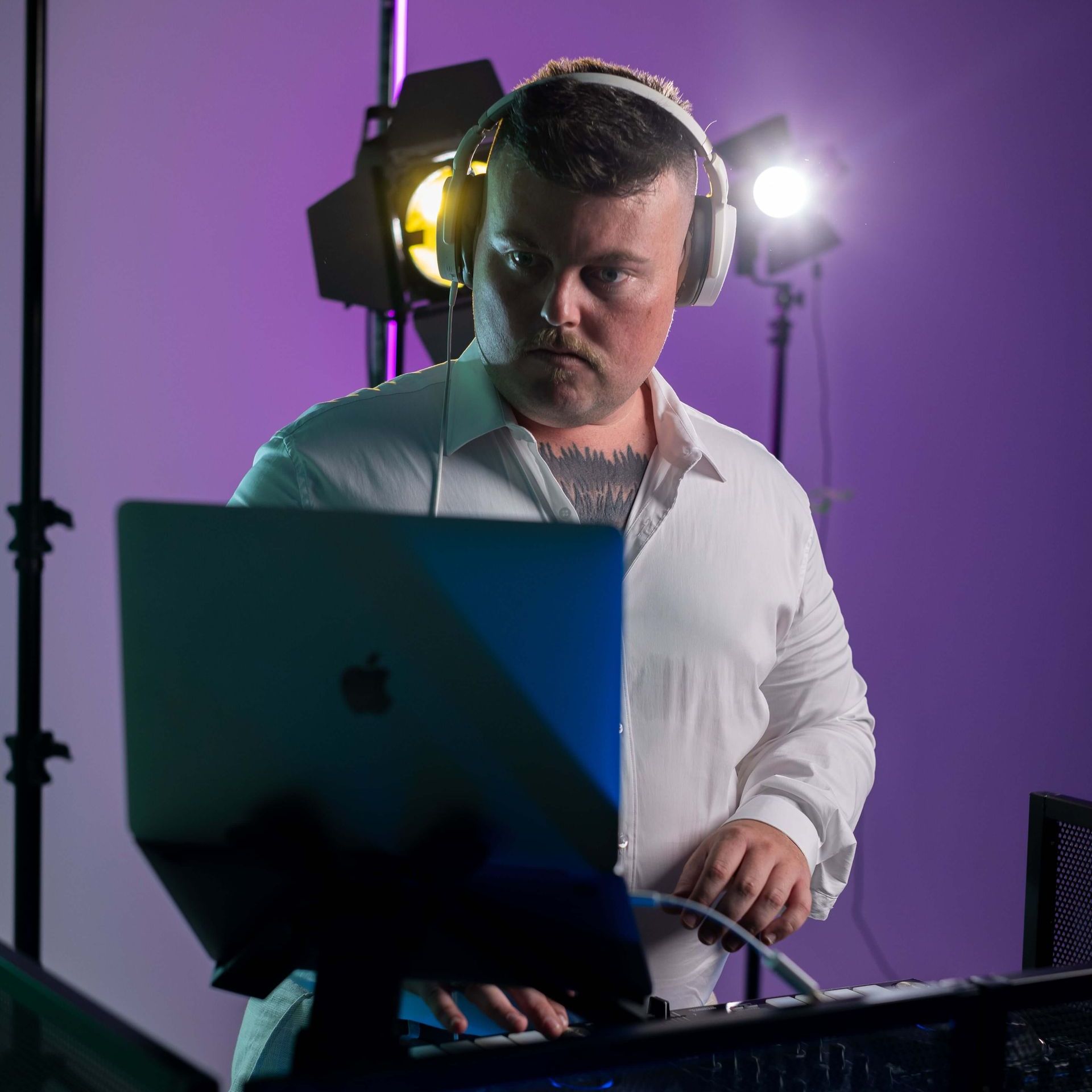 DJ in a white shirt wearing headphones, working on a laptop with studio lighting in a purple-lit room.