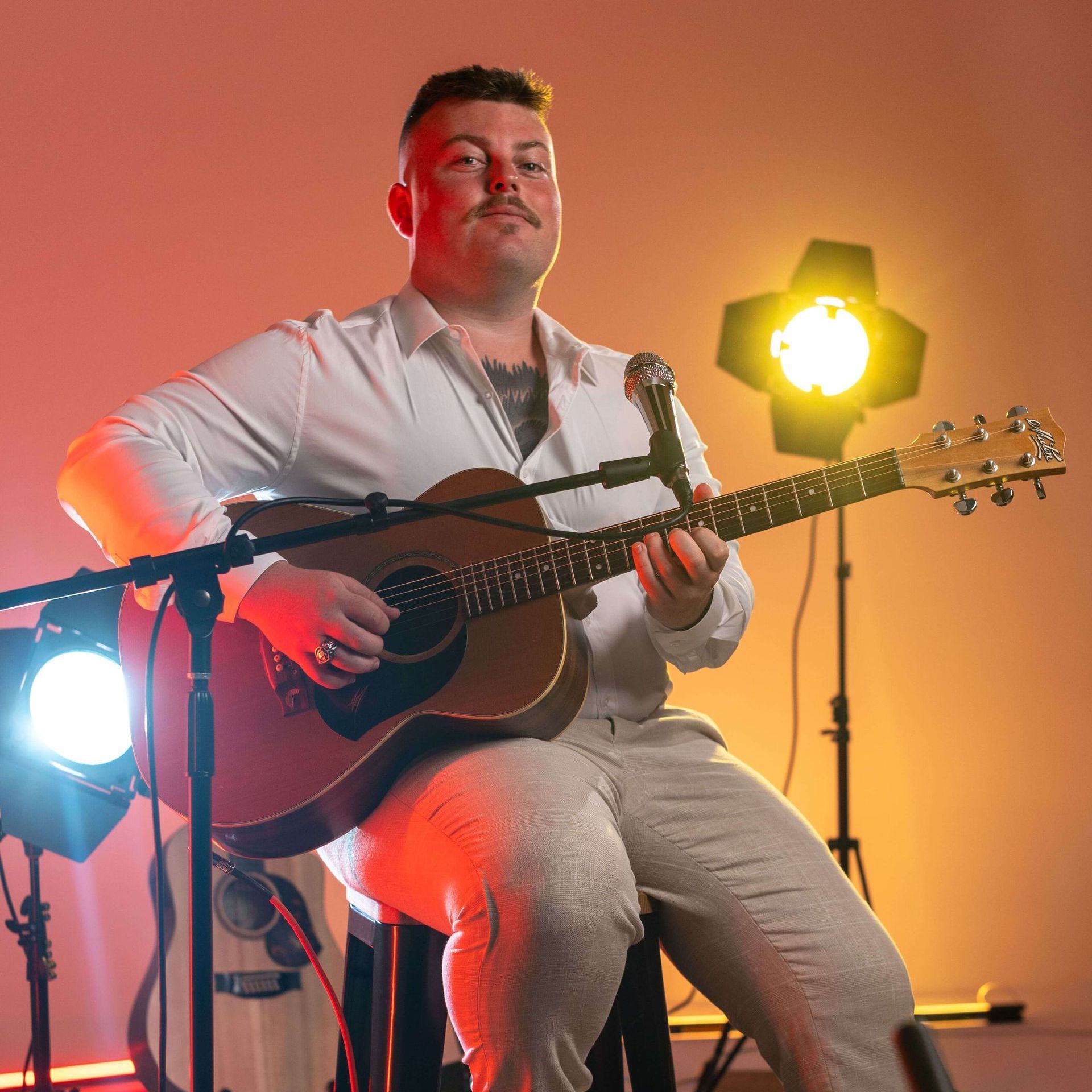 A man with a mustache playing an acoustic guitar, seated, under studio lights.