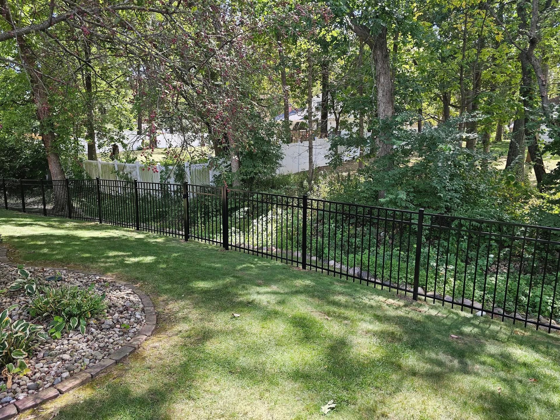 Black metal fence encloses a green lawn, trees in the background, sunny day.