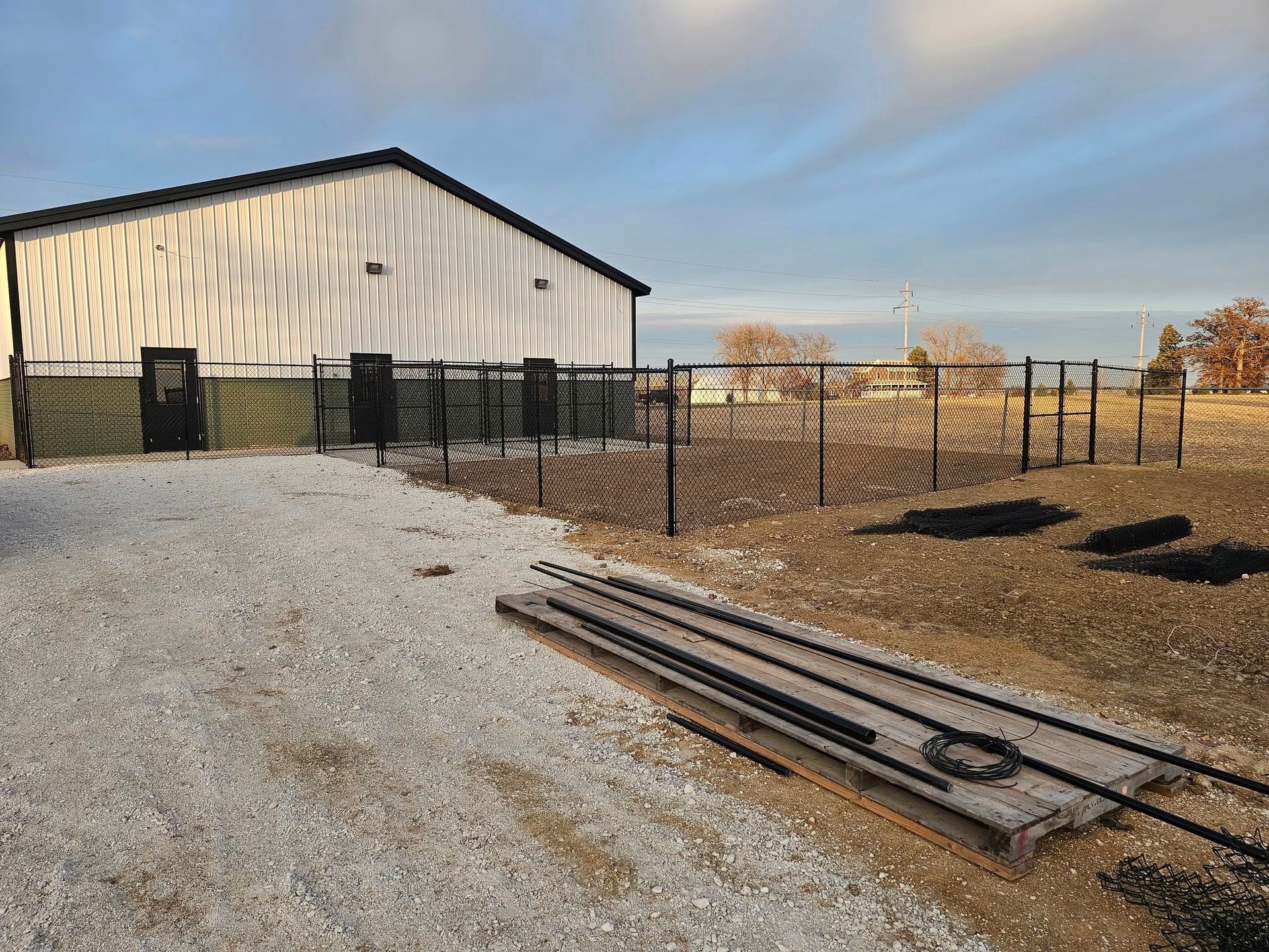 Black fence surrounds a gravel area next to a white industrial building. Steel fence components sit in foreground.