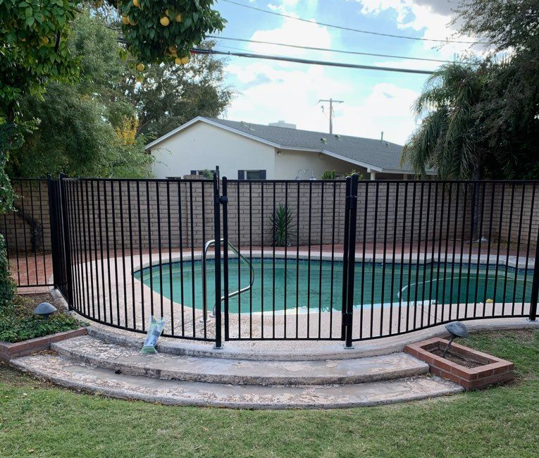 Black metal fence encloses a kidney-shaped swimming pool in a backyard. A brick wall and house are in the background.