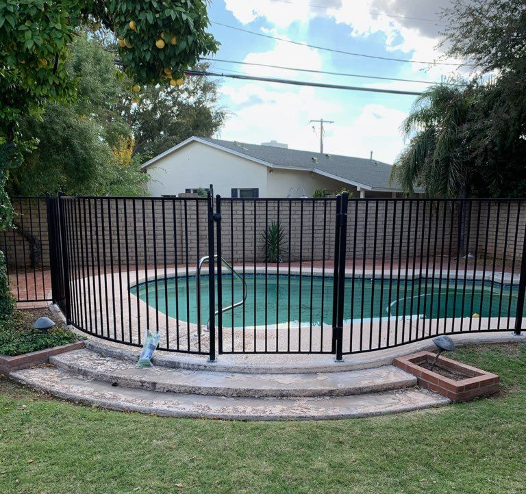 Black fence surrounds a pool with brick steps, green lawn, and a house in the background.
