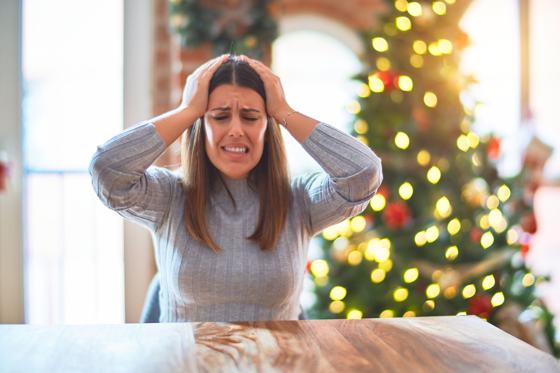 Woman with hands on head, looking stressed, in front of a decorated Christmas tree.
